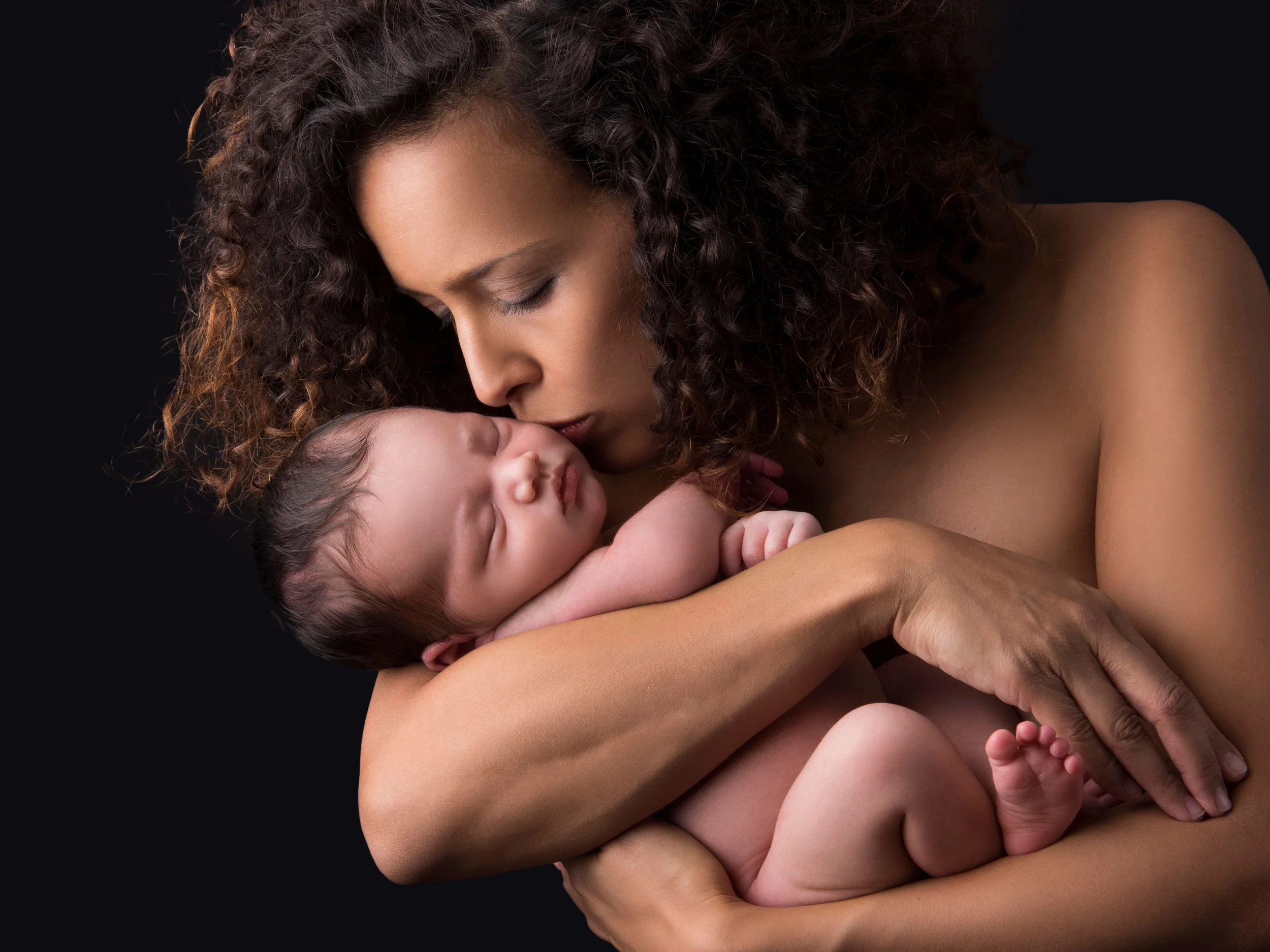 A woman with curly hair holding and cuddling a sleeping baby against a dark background.