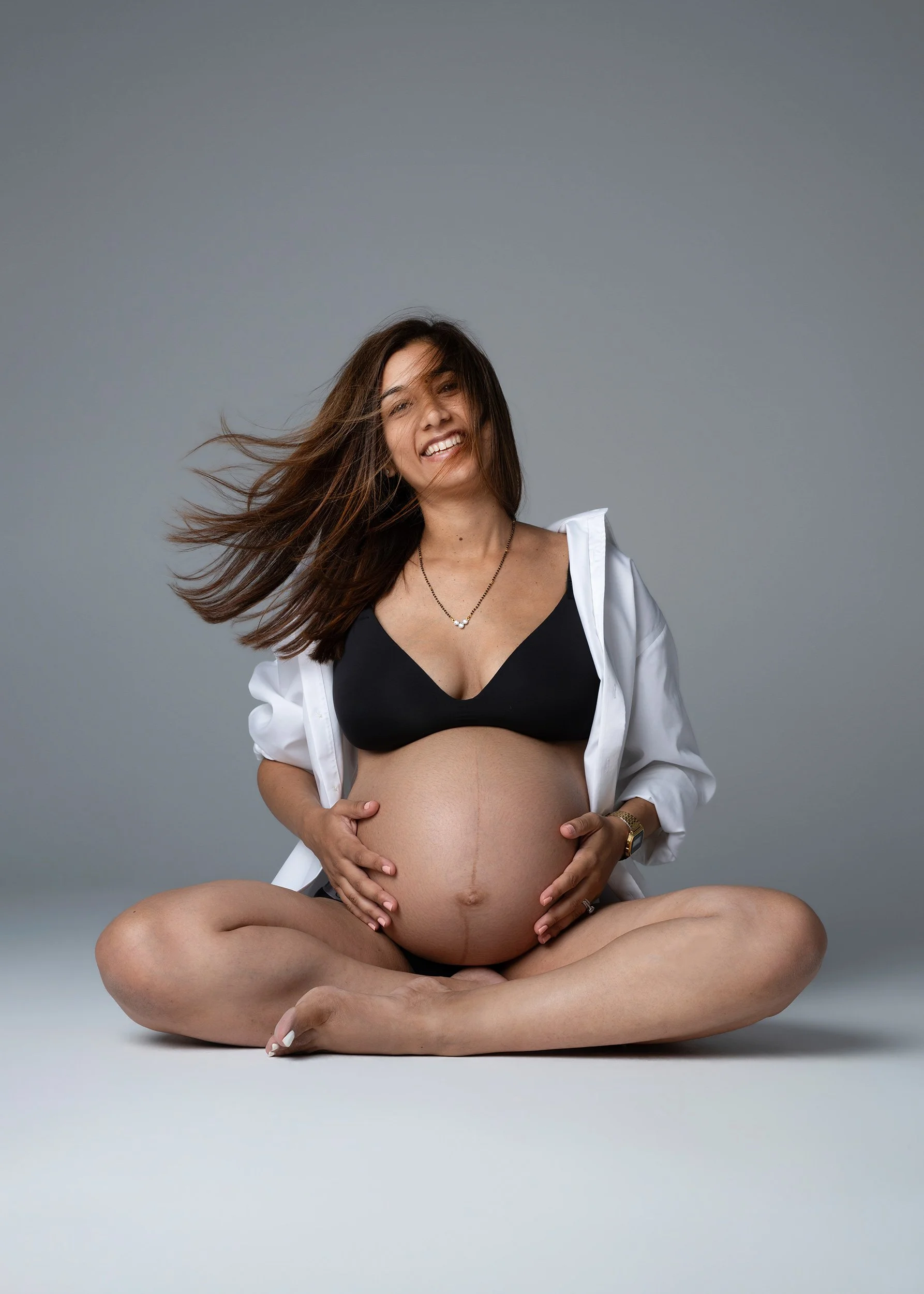 Pregnant woman seated in a white shirt and black bra during a modern London maternity photoshoot.