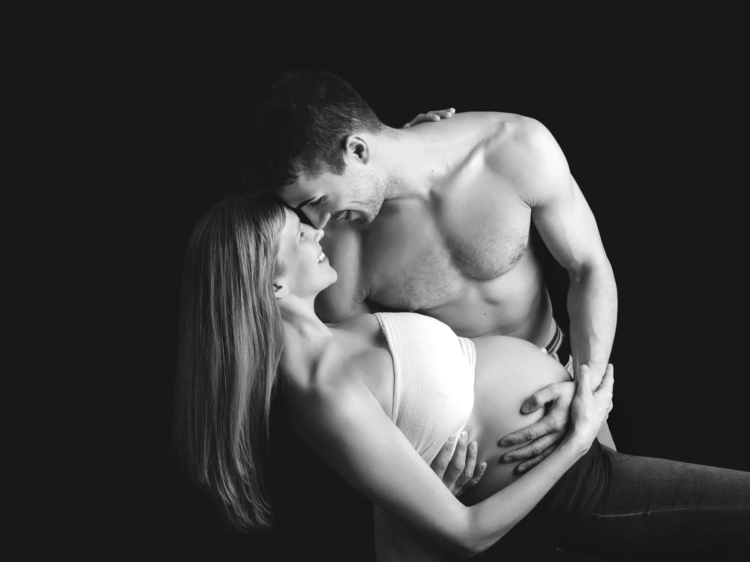 Black-and-white couples maternity portrait of an expectant mother embracing her shirtless partner, smiling as they gently touch foreheads in the studio.