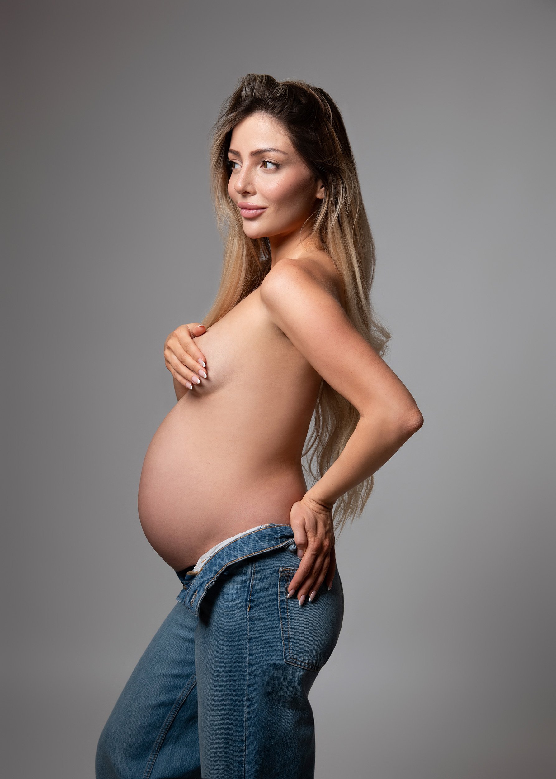 A pregnant woman with long brown hair, wearing jeans, partially topless, posed against a plain gray background.