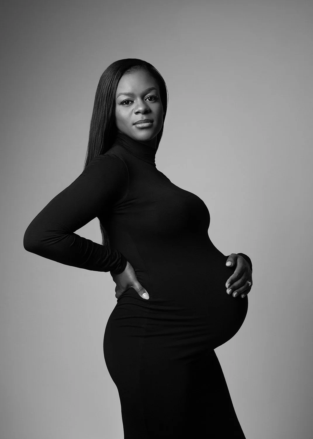 Black and white maternity studio portrait of a woman wearing a long black turtle neck dress.