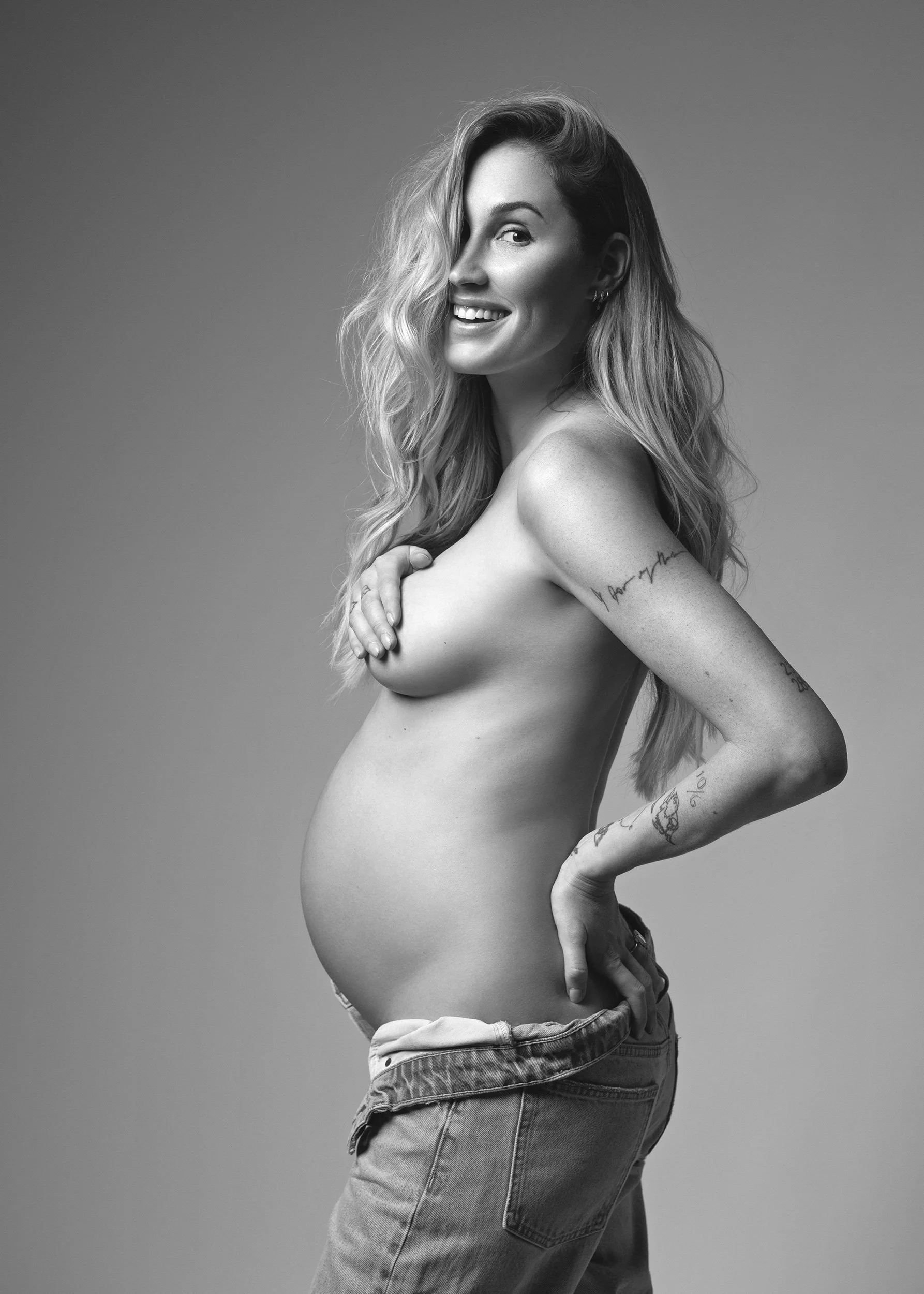 Black-and-white maternity portrait of a smiling pregnant woman with long wavy hair and arm tattoos, wearing jeans and covering her chest while holding her belly against a plain studio background.