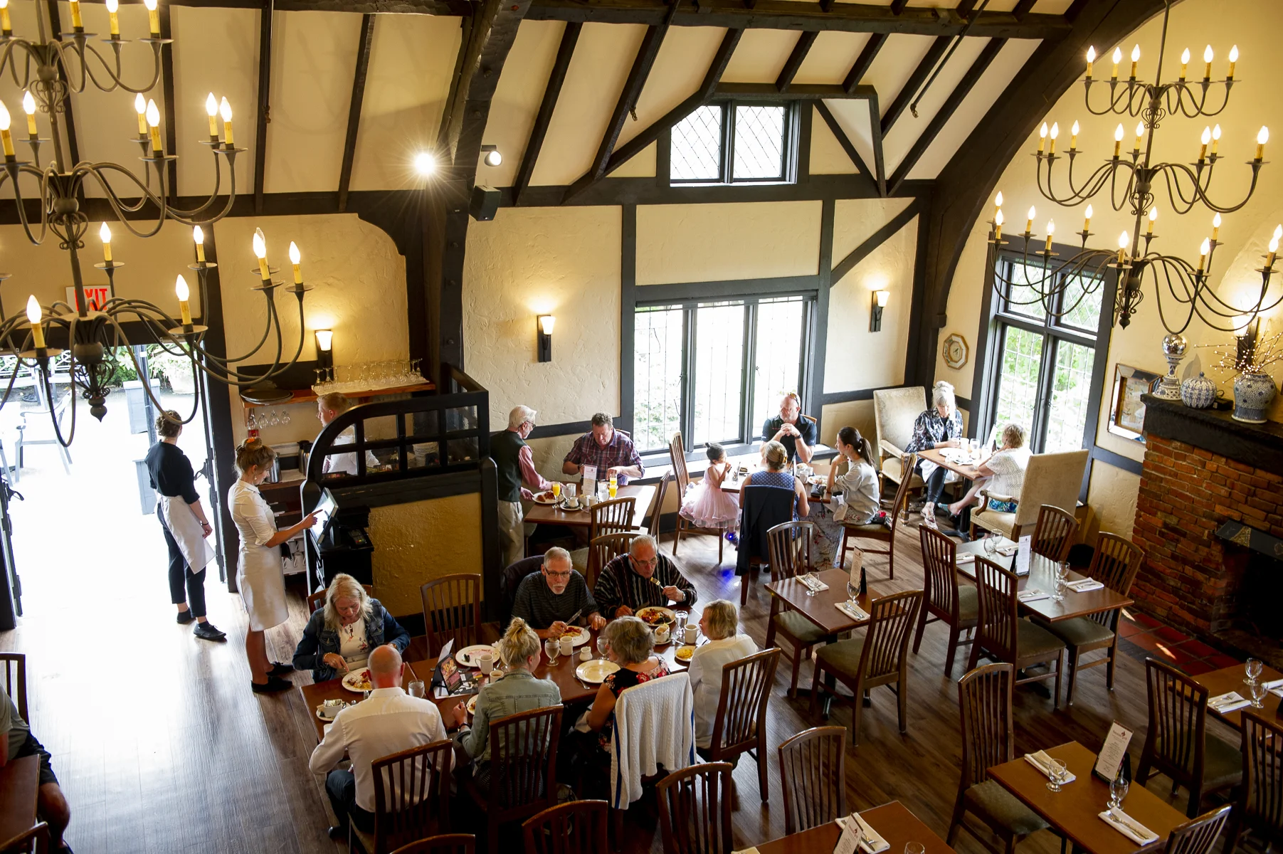 A cozy restaurant interior with high vaulted ceilings and exposed dark wooden beams. Chandeliers with candle-like light bulbs hang from the ceiling. Large windows allow natural light inside. Guests are dining at wooden tables, with some kids and families present. Waitstaff serve food, and a waitress reads a menu or device near the entrance.