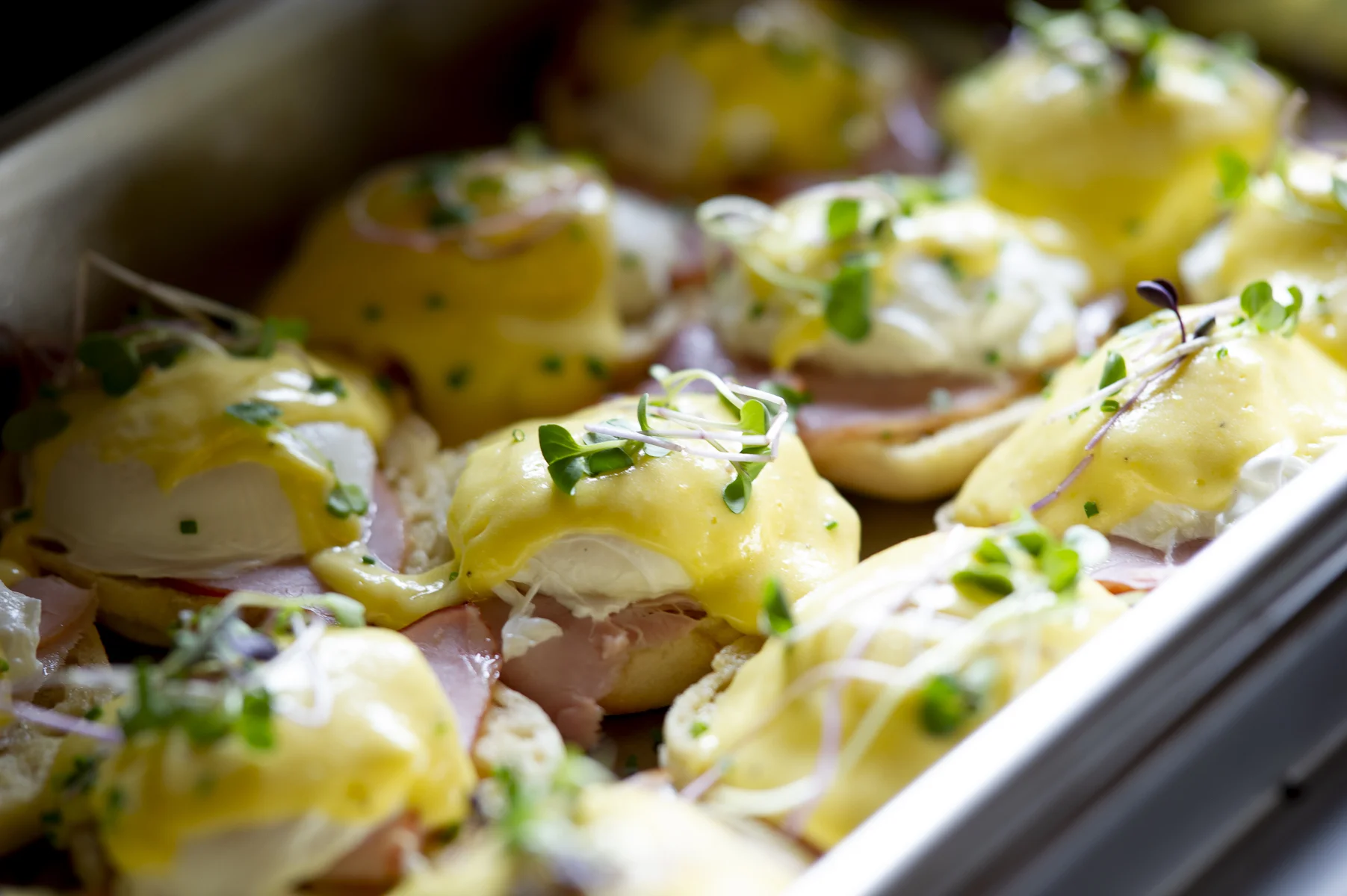 Close-up of eggs Benedict with slices of ham, topped with hollandaise sauce and microgreens on an eggs Benedict platter.