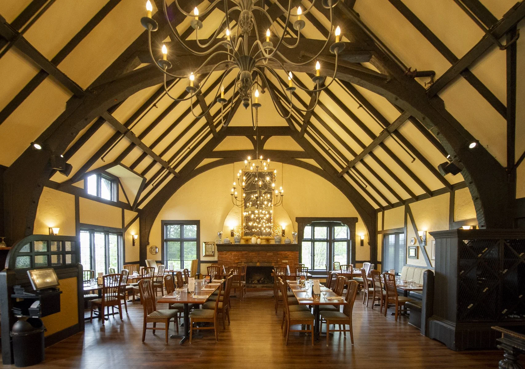 A picture of the interior of The Fireside Room shows a rustic dining room with high vaulted ceiling, hanging chandeliers, wooden tables and chairs, a brick fireplace in the background, and large windows allowing natural light.