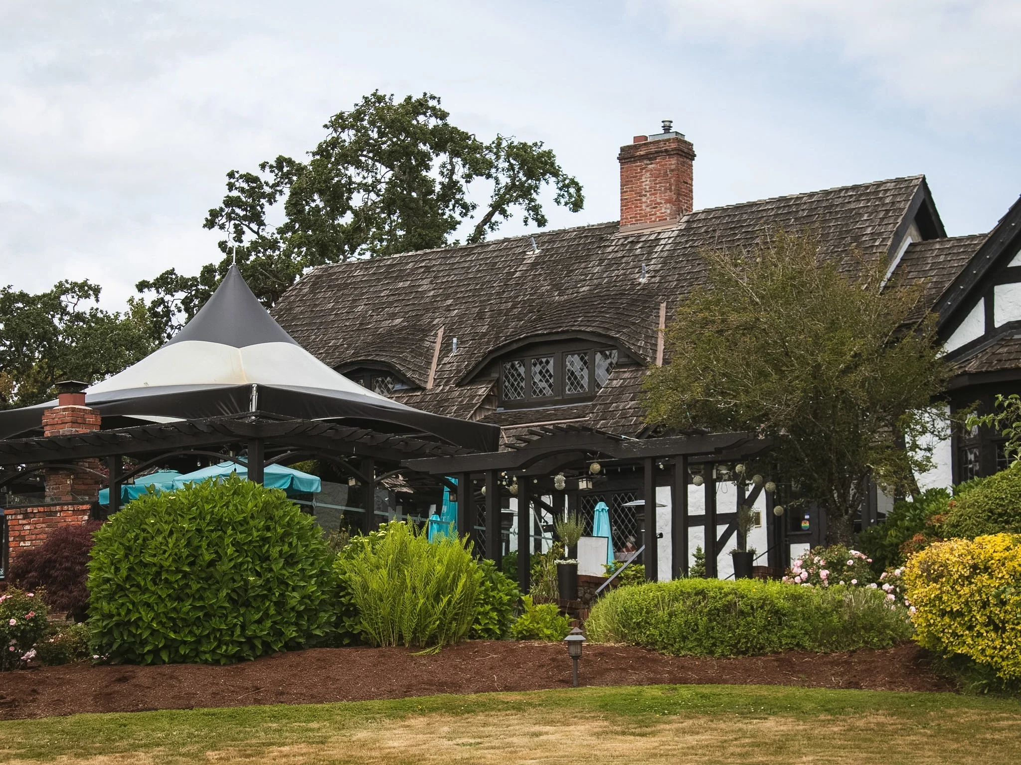 A historic house with a steeply sloped roof, brick chimney, and decorative windows, surrounded by a garden with lush green bushes, colorful flowers, and outdoor patio seating under umbrellas.