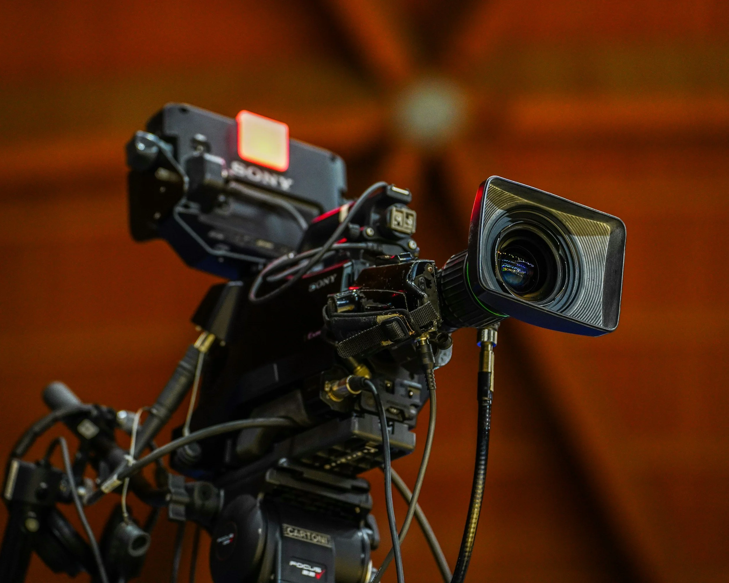 Close-up of a professional video camera with various electronic components and wiring, set against a blurred brown background.