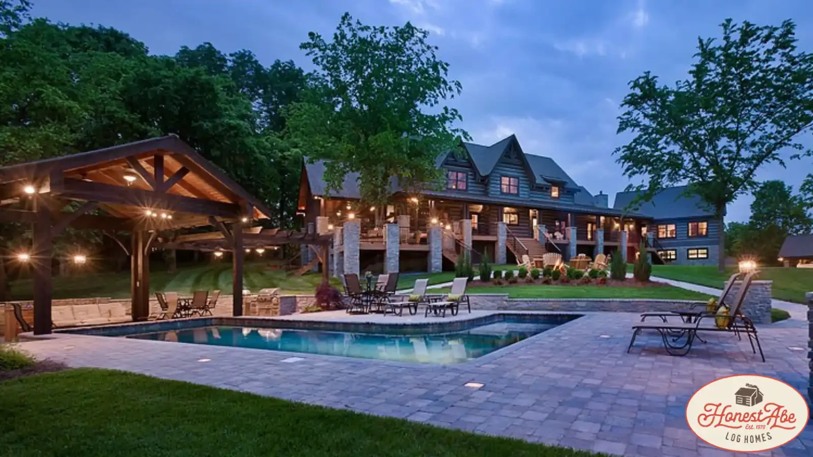 Rear Exterior view of a lakefront Log and Timber Frame Cabin with Stone Paver Patio, In-Ground Pool and Custom Framed Cabana, full length wrap around deck at dusk, home design by Honest Abe Cabins and BuildBuyRefi.com financing.