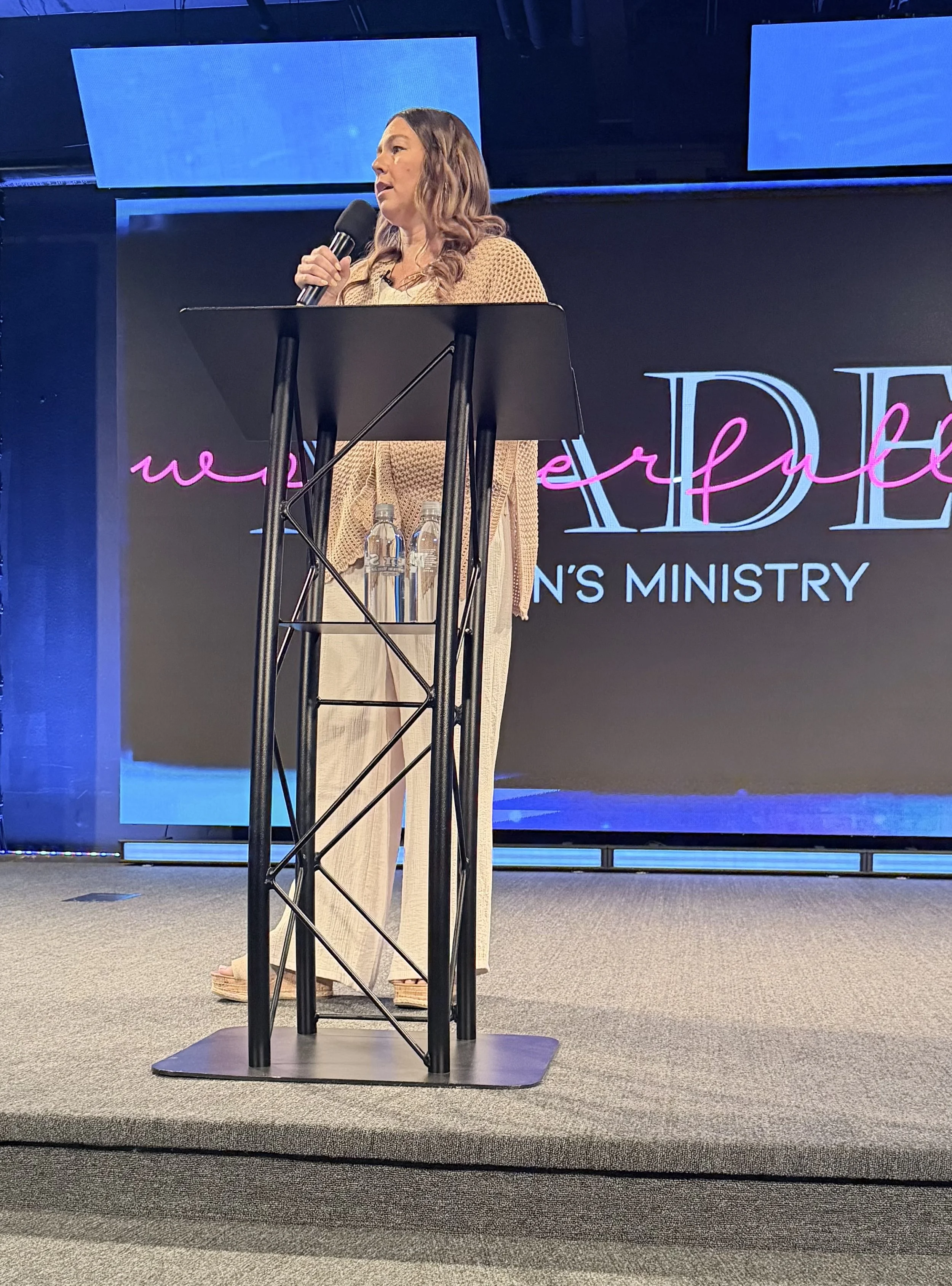 A woman standing at a podium, speaking into a microphone, with a large screen behind her displaying partially visible text that reads 'women' and 'ministry'.