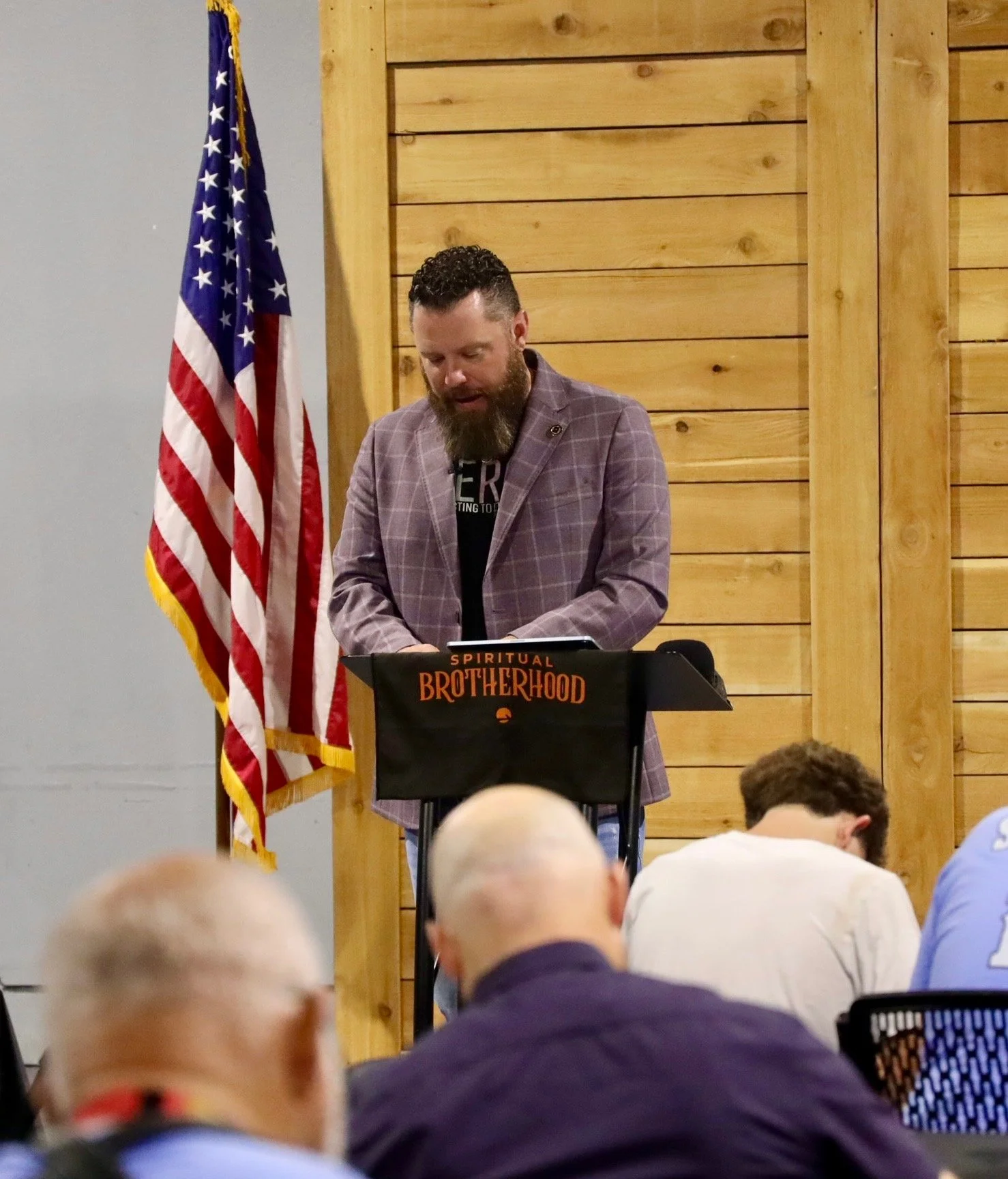 A man with a beard and a checkered blazer is standing at a lectern with a sign that says 'Spiritual Brotherhood'. He is in front of an American flag and a wooden wall, speaking to an audience in a room with some people visible in the foreground.