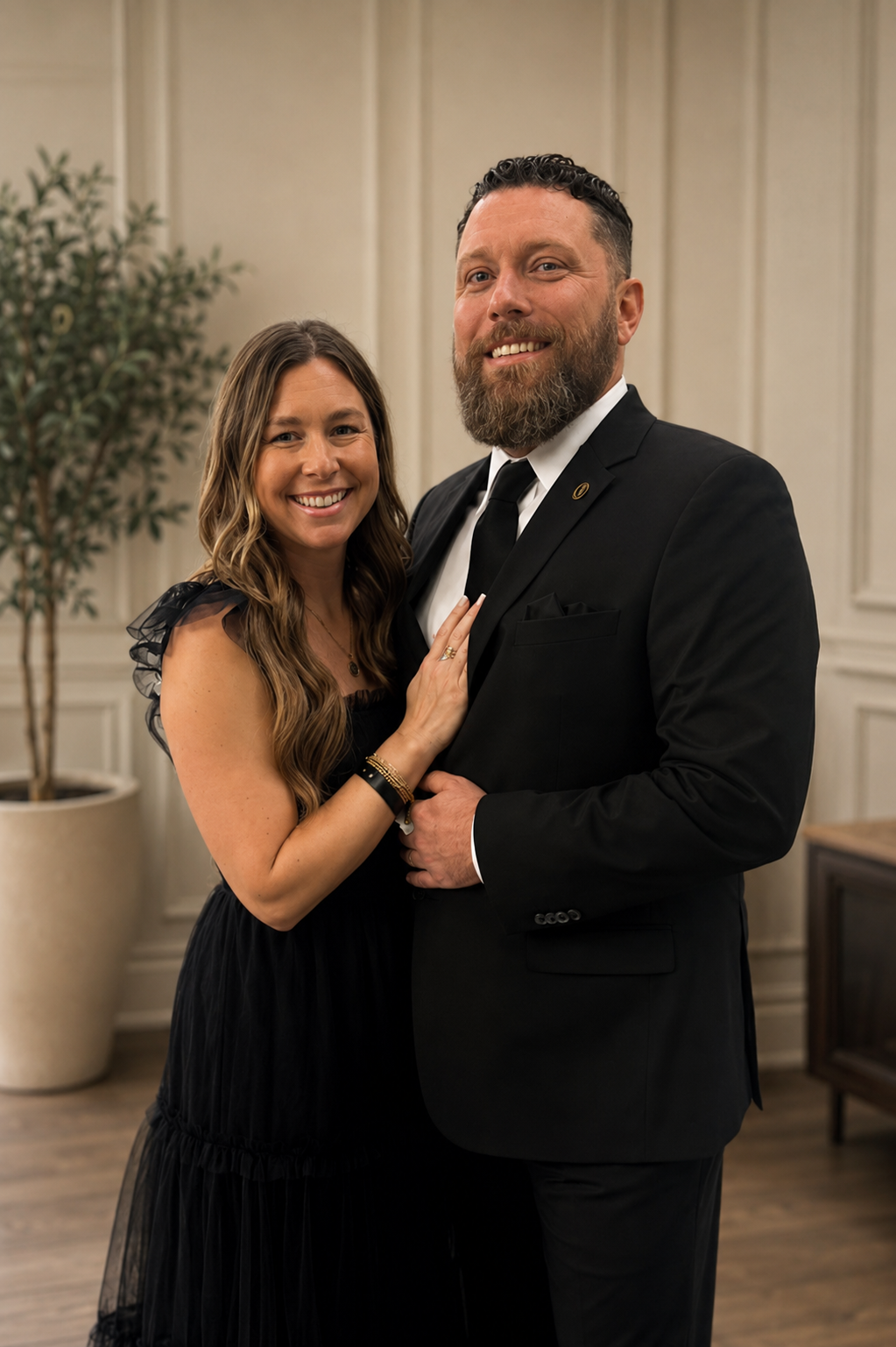 A smiling woman and man dressed in formal attire, standing closely together in an elegant room. The woman has long wavy brown hair, wearing a black dress with ruffled sleeves, and the man has short dark hair and a beard, wearing a black suit with a tie. They are indoors with cream-colored walls, a potted plant, and wooden furniture in the background.