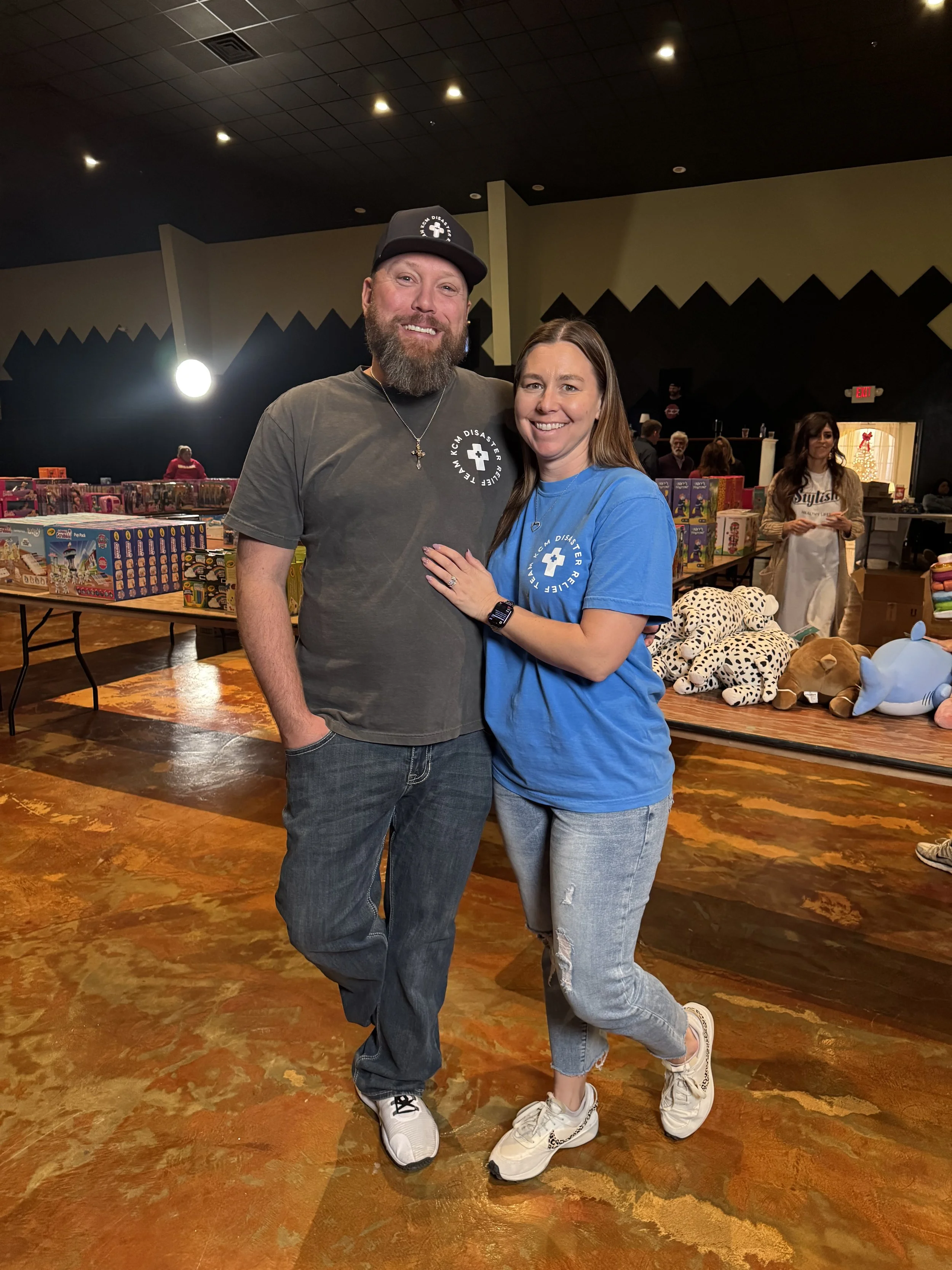 A man with a beard and a woman with long hair are posing together at a charity event, with tables of toys and stuffed animals behind them. The man is wearing a black cap and a dark T-shirt, while the woman is wearing a blue T-shirt, jeans, and white sneakers. There are other people and a Christmas tree in the background.