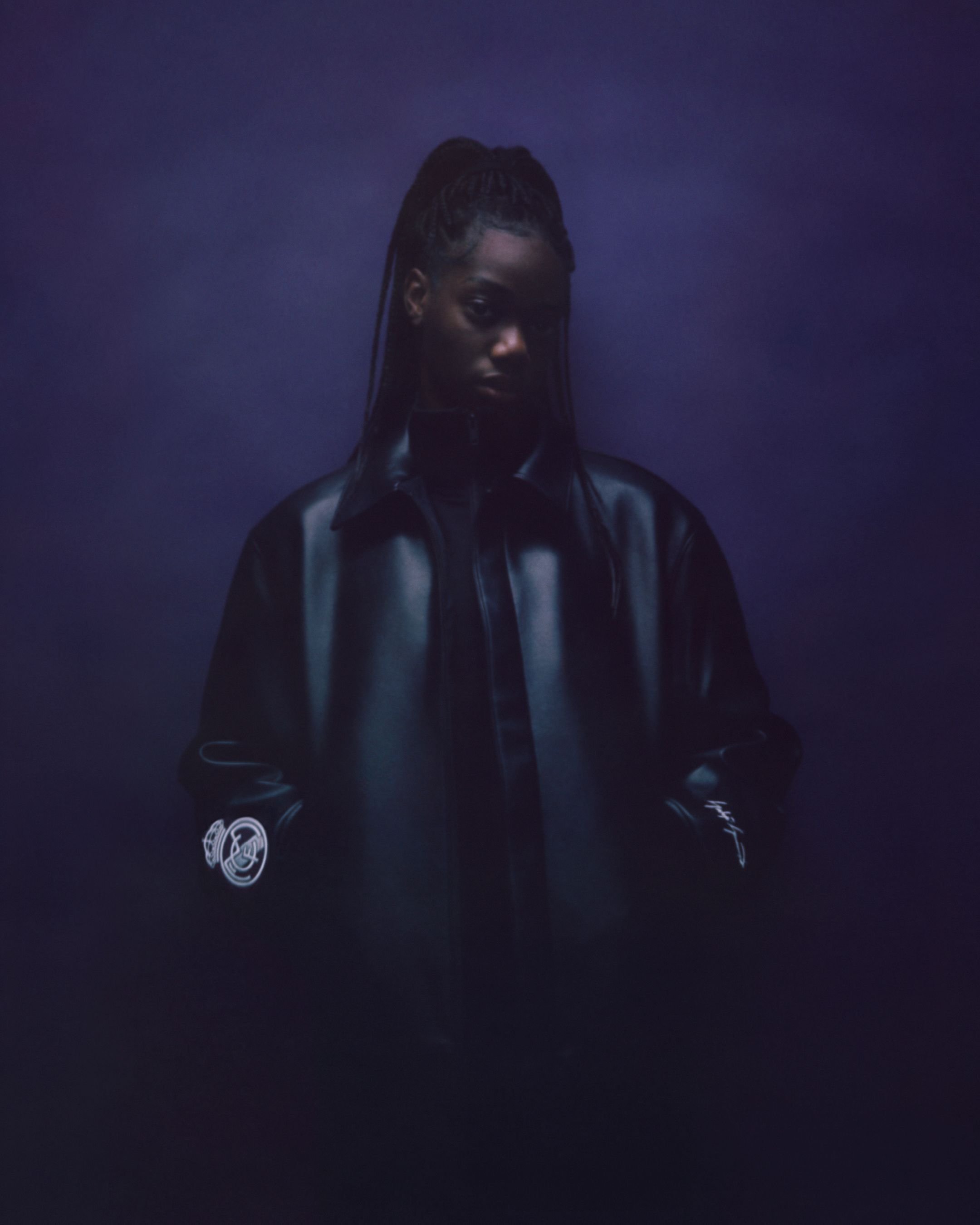 A woman with dark skin and dreadlocks wearing a black leather jacket, standing against a dark purple background.