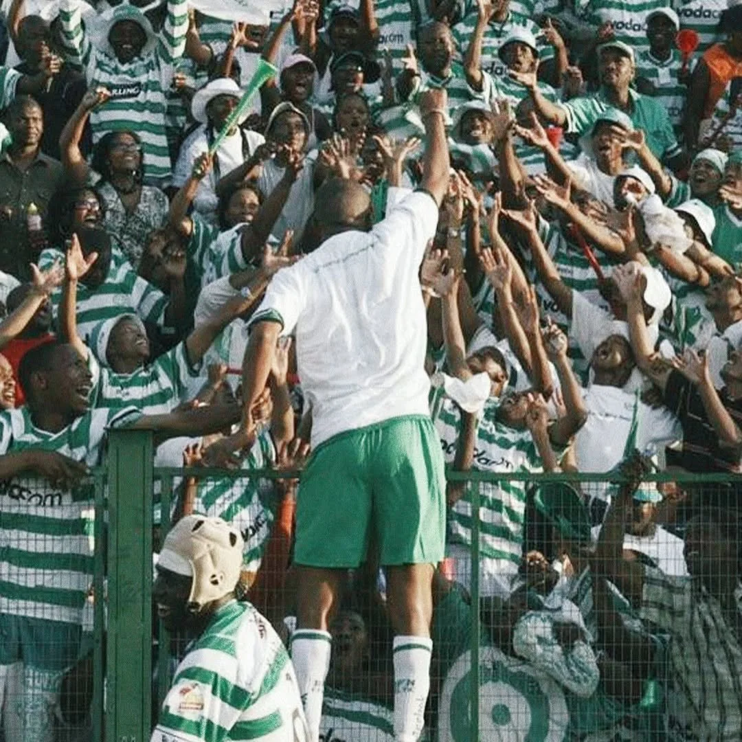 A group of football fans dressed in green and white striped jerseys celebrate, with one in the center reaching out to a man on a ledge.