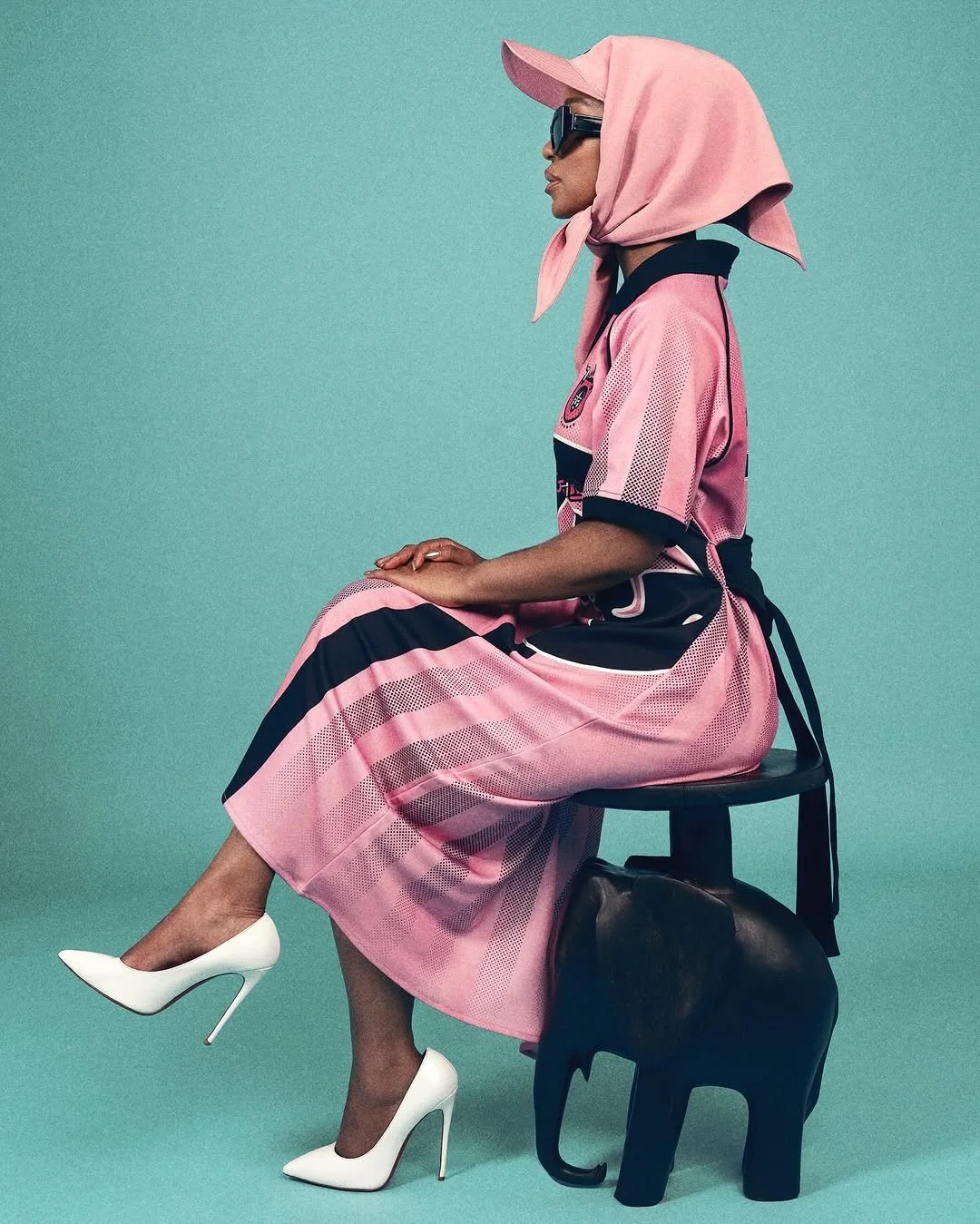 A woman dressed in a pink and black sports jersey, pink headscarf, and white high heels, sitting on a black stool shaped like an elephant against a teal background, wearing sunglasses.