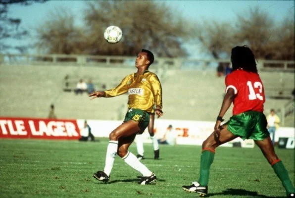 Two soccer players, one in yellow and one in red, compete on a grass field with a soccer ball in mid-air during a match.