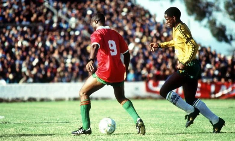 Two soccer players competing for the ball during a match with a crowd of spectators in the background.