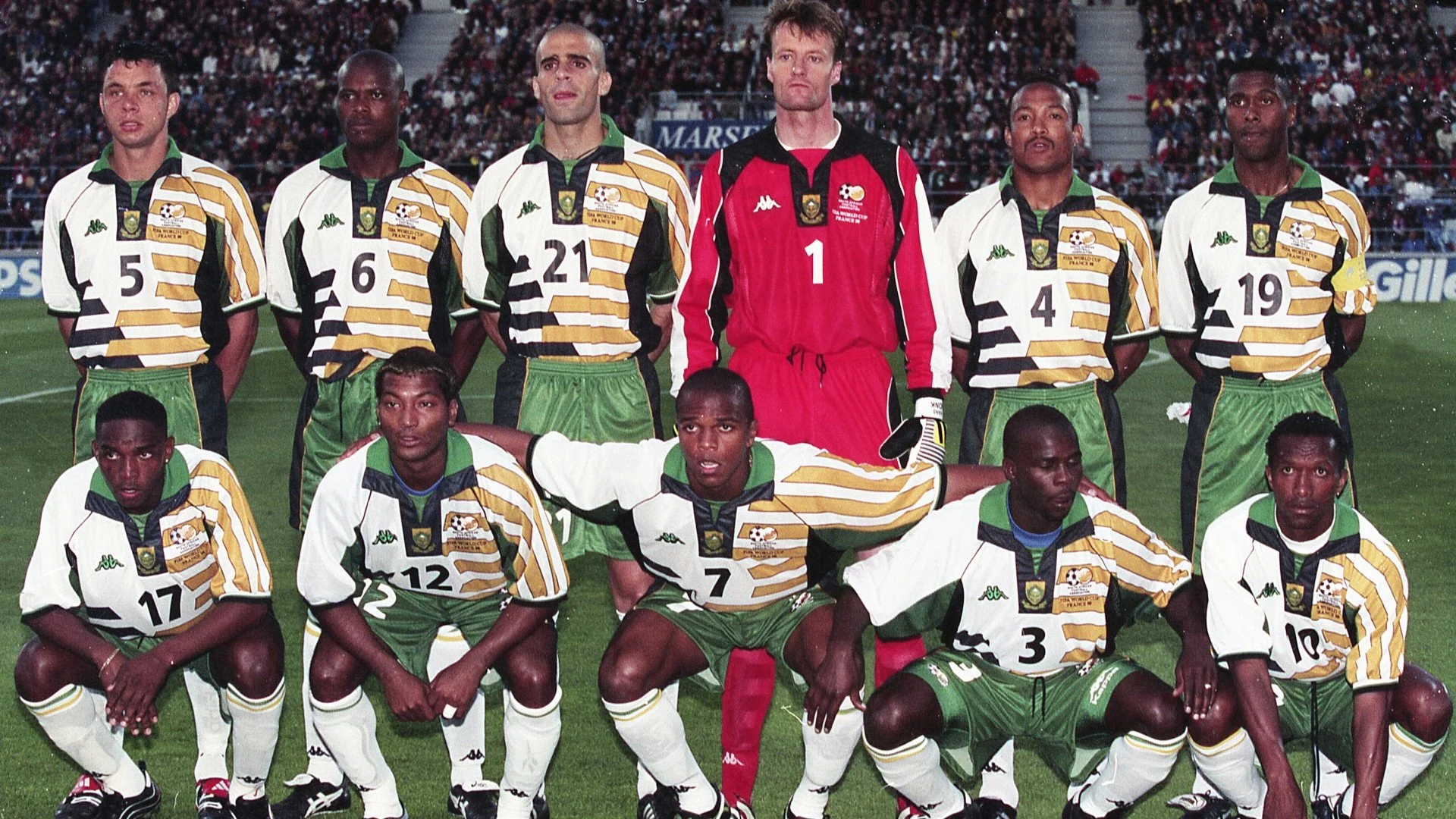 Soccer team in uniform on the field, posing for a team photo before a match, with a stadium and spectators in the background.