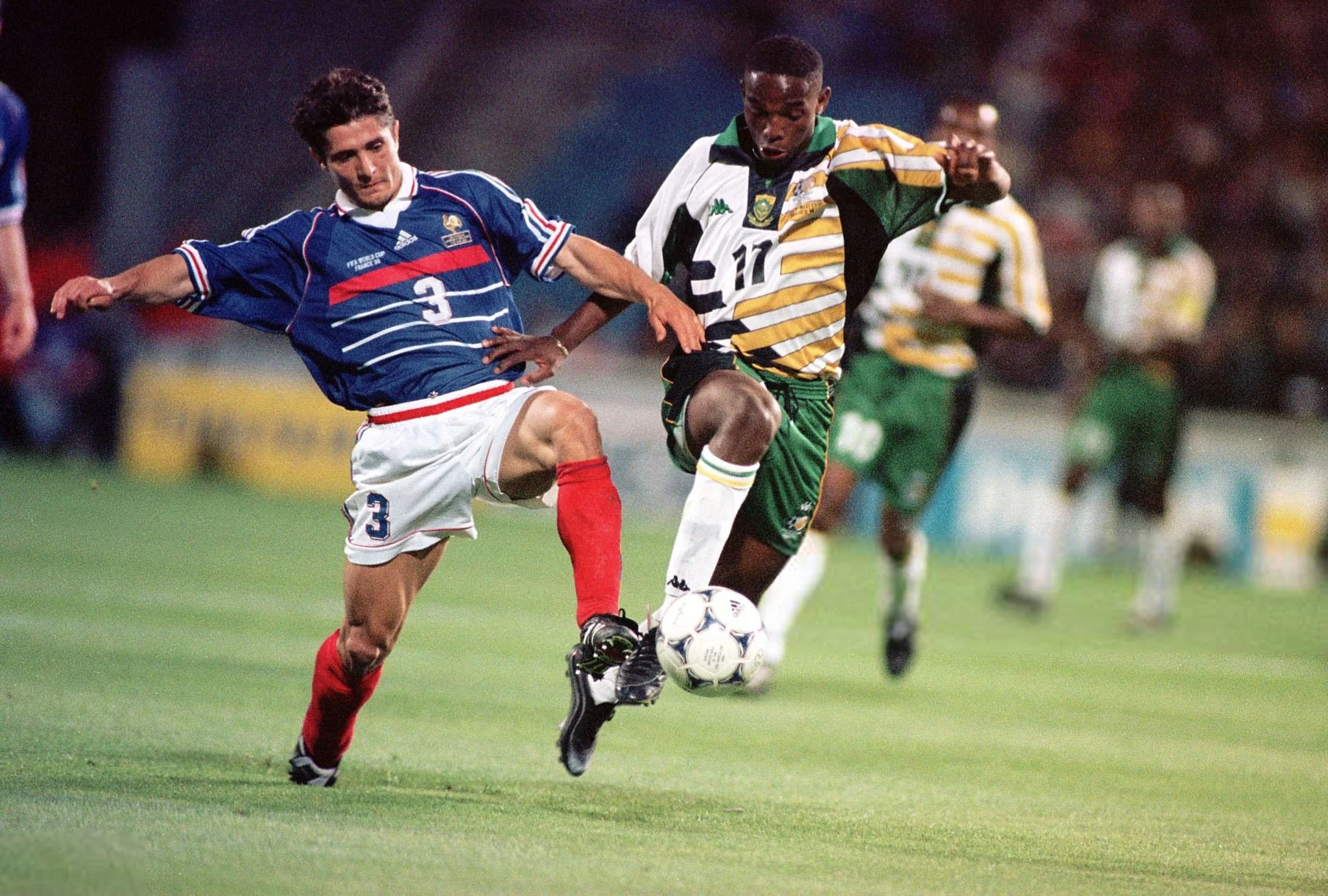 Two soccer players, one in a blue jersey and the other in a green and yellow jersey, compete for the ball during a match on a grassy field, with other players in the background.