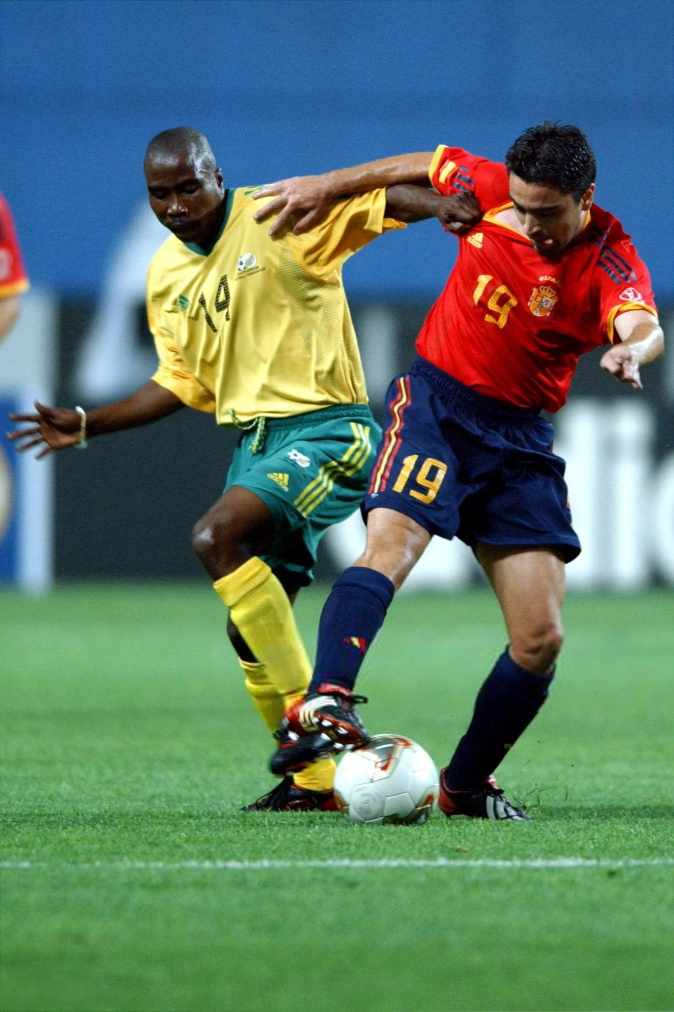 Two soccer players competing for the ball, one wearing a yellow and green uniform and the other in a red and navy blue uniform, on a green soccer field.