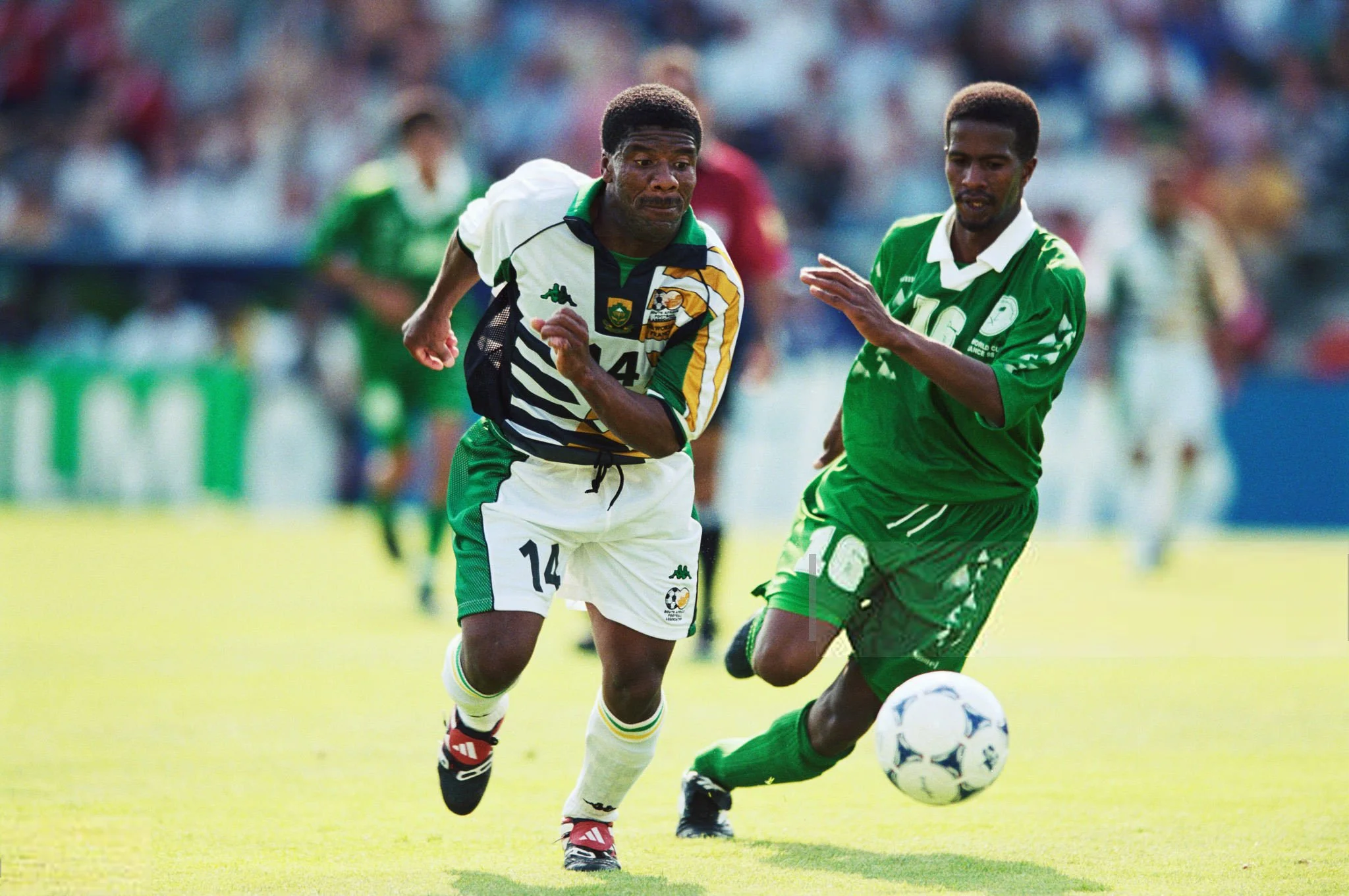 Two soccer players competing for the ball during a match, with one wearing a white and black jersey and the other in a green jersey, on a grassy field with spectators in the background.