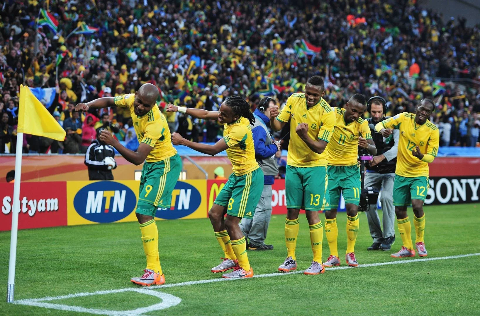 A group of five soccer players in yellow and green team uniforms celebrating on the sideline of a soccer field, with a large crowd of spectators in the background.
