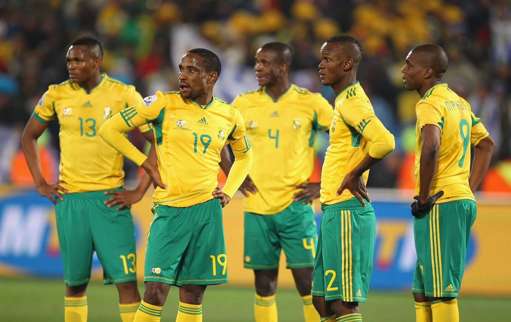 Five male soccer players dressed in yellow and green uniforms, standing on a field during a match, with animated crowd in the background.