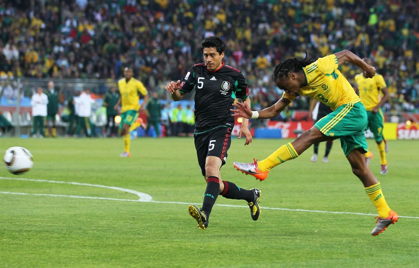 Two soccer players, one in a black jersey with the number 5 and another in a yellow jersey, are competing for the ball during a match on a well-kept grass field, with a large crowd of spectators in the stands.