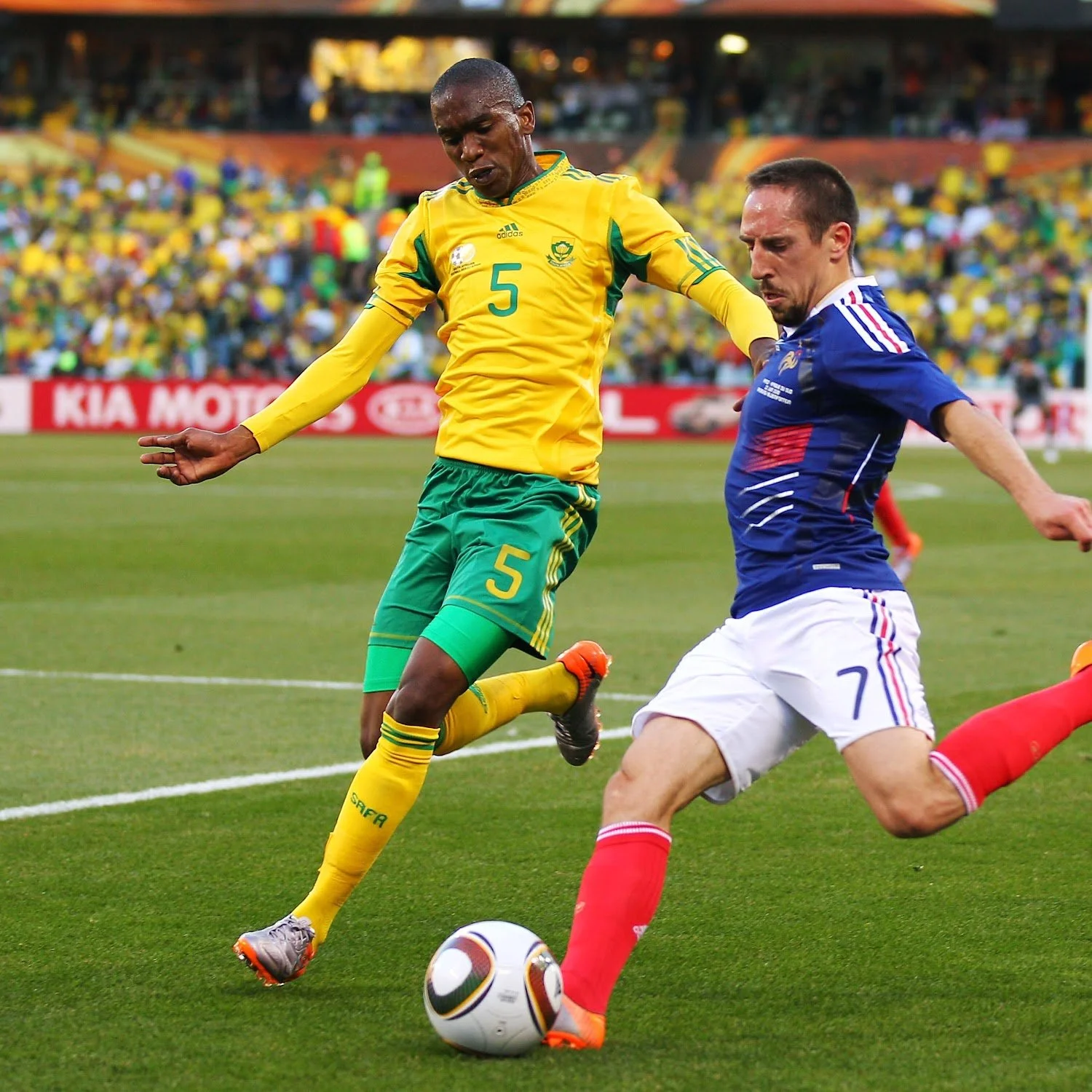 Two soccer players from opposing teams compete for the ball during a match on the field. The player in yellow and green is wearing jersey number 5, and the player in blue, white, and red is wearing jersey number 7.