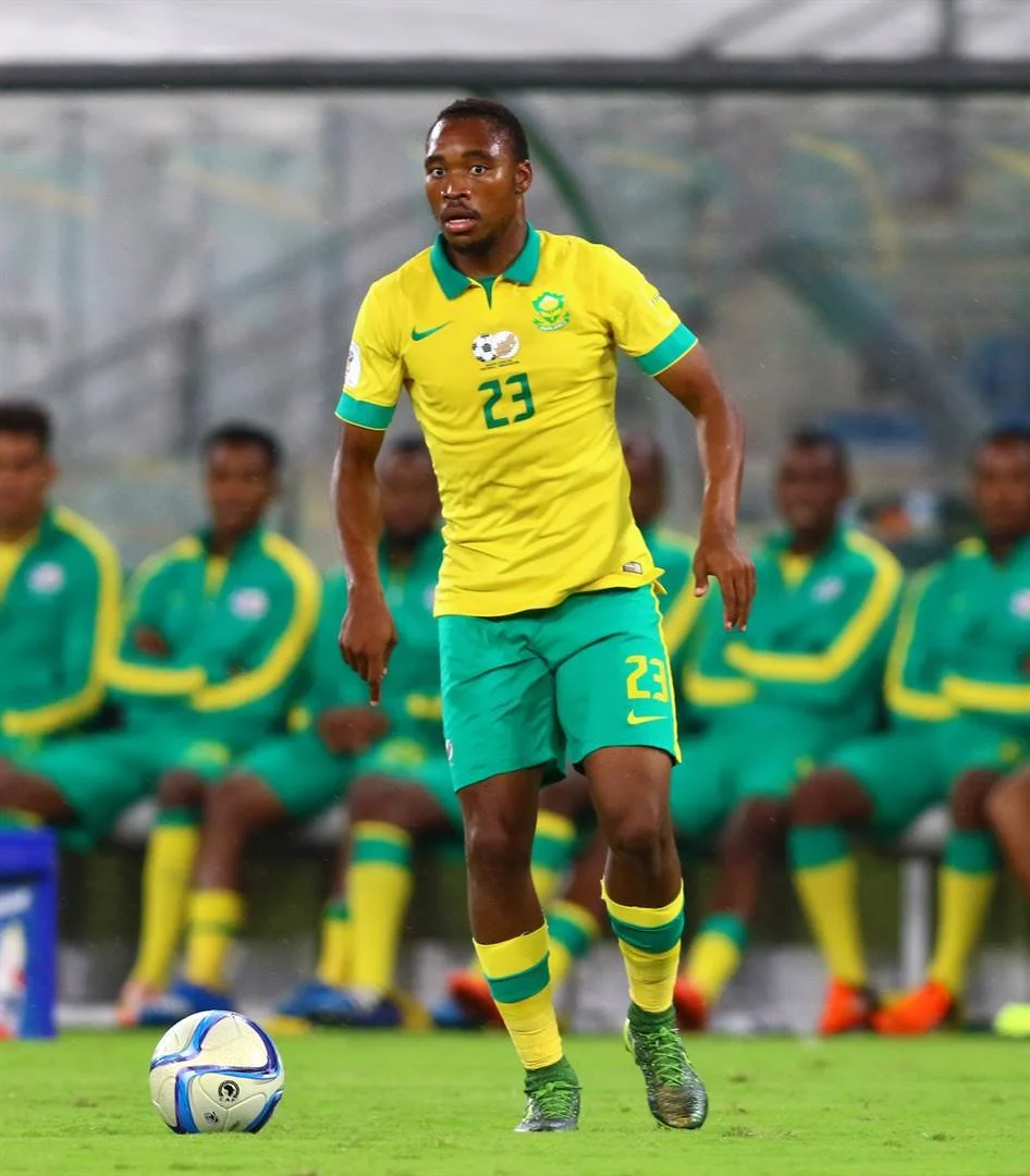 A soccer player wearing a yellow and green uniform with the number 23 is about to kick a soccer ball on the field. Several teammates in matching uniforms sit on the bench in the background.
