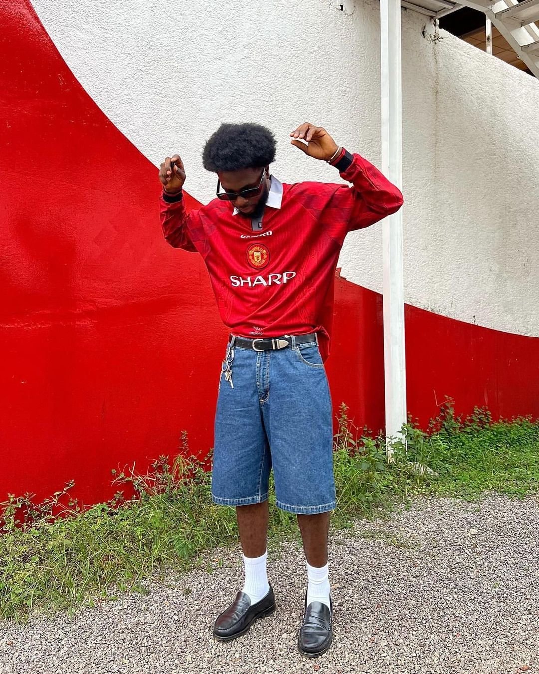 A young man with an afro hairstyle wearing sunglasses, a red soccer jersey with the Manchester United logo and 'Sharp' sponsor, denim shorts, white socks, and black loafers, standing outdoors with a red and white wall background.