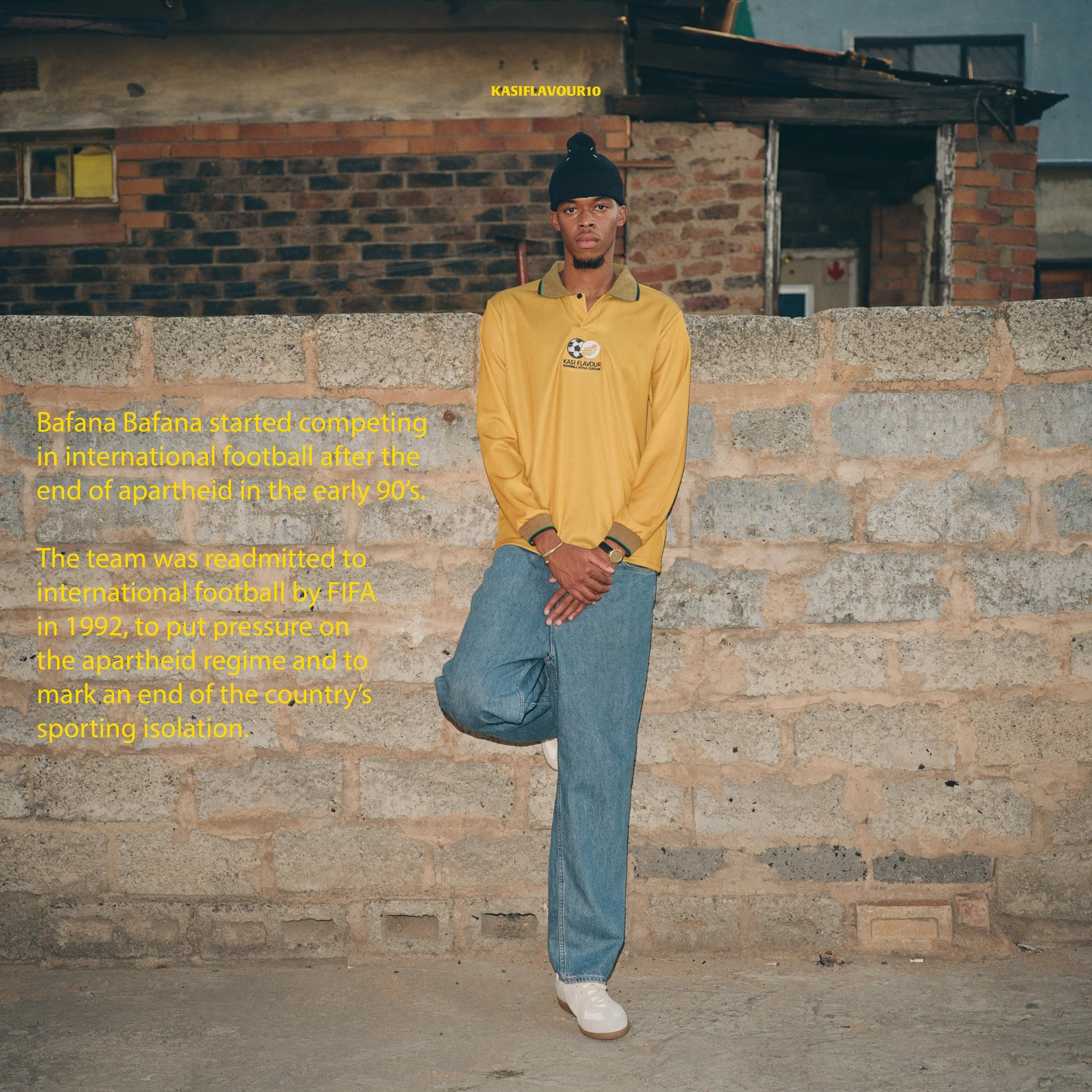 A young man wearing a yellow long-sleeve shirt, blue jeans, white sneakers, and a black beanie stands against a brick wall in an outdoor setting, with a message about the history of Bafana Bafana soccer team and apartheid.
