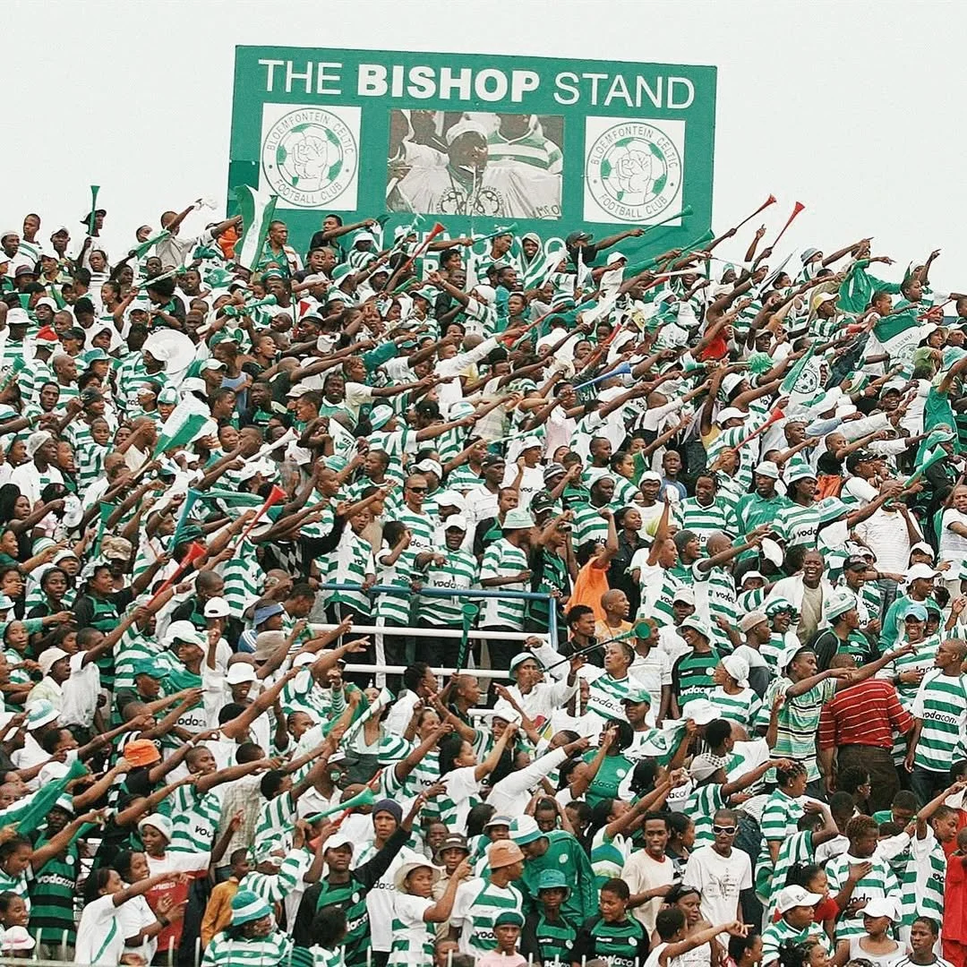 Crowd of football fans wearing green and white striped shirts, sitting in a stadium, raising their arms in cheer, in front of a large sign that reads "The Bishop Stand."