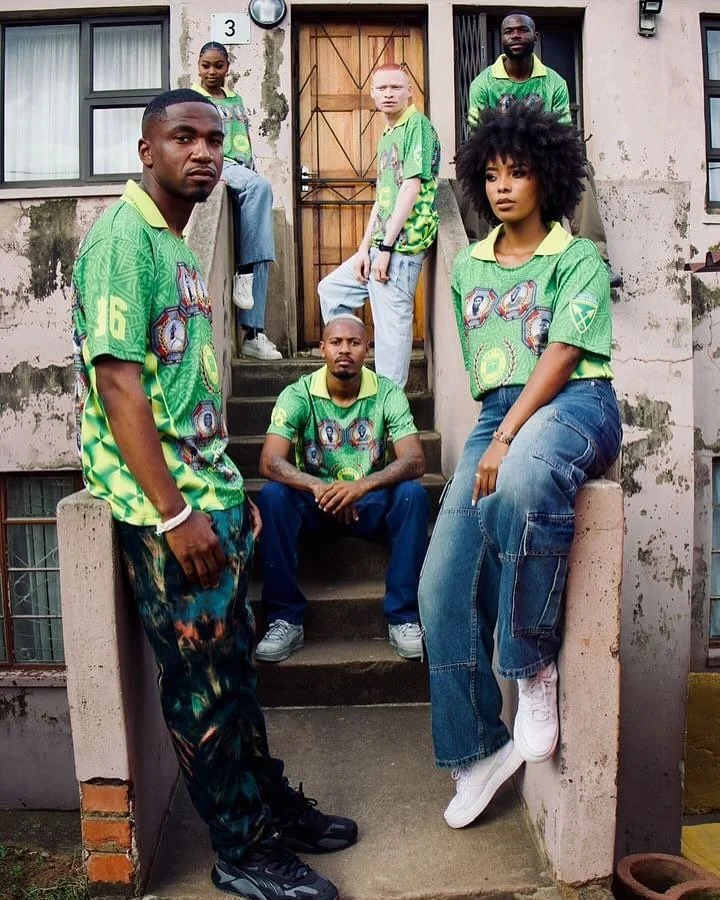 Six diverse young adults, four men and two women, posing on the steps outside a building with a weathered wall and windows. They are dressed in matching green shirts with colorful designs.