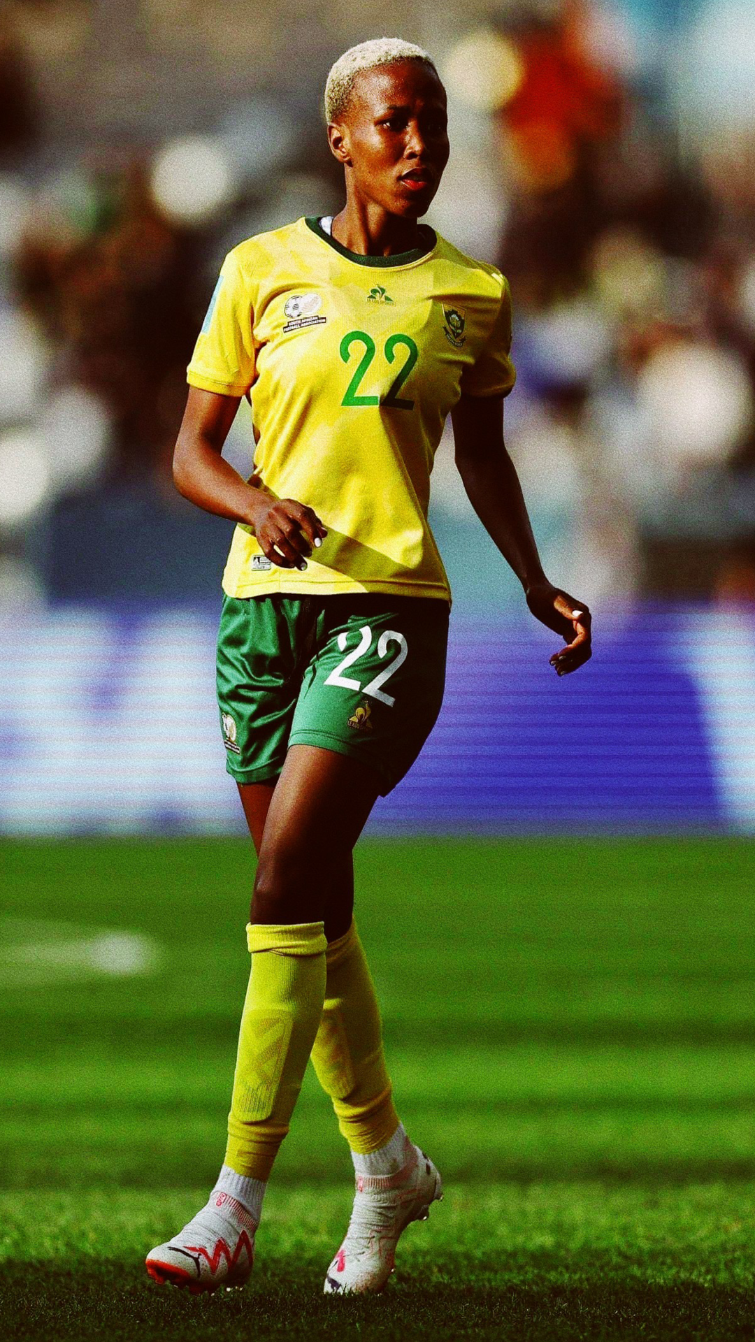 A female soccer player with a dark skin tone and short, bleached blonde hair is on the field wearing a yellow and green uniform with the number 22. She appears to be in motion during a game.