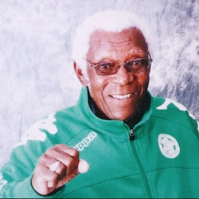 An elderly man wearing glasses and a green jacket, smiling and making a fist with his right hand.