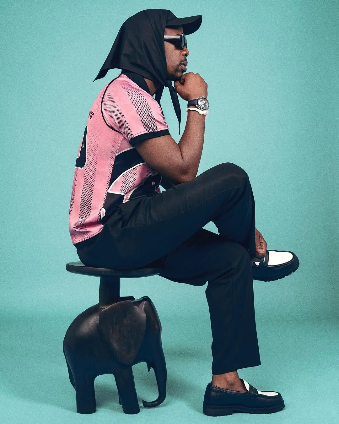 A man sitting on a stool designed like an elephant against a light teal background. He is wearing a pink and black sporty shirt, black pants, black and white shoes, sunglasses, and has a black hat with a cloth covering his head. He is resting his chin on his hand and appears to be deep in thought.