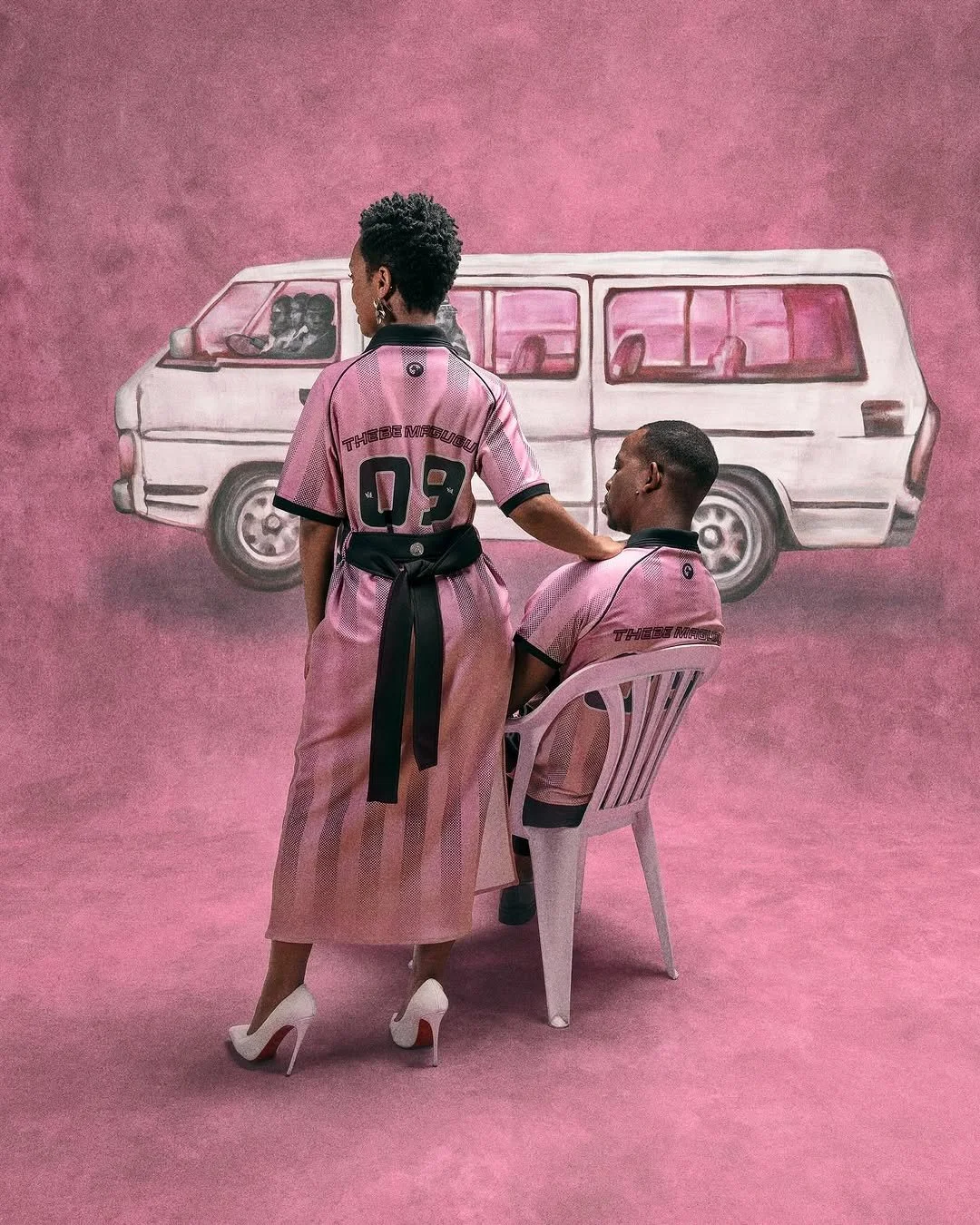 An art installation features two models in pink sports jerseys with black accents in front of a pink painted background with a mural of a white van. One model is standing, and the other is seated on a white plastic chair, with the standing model's hand on the seated model's shoulder.