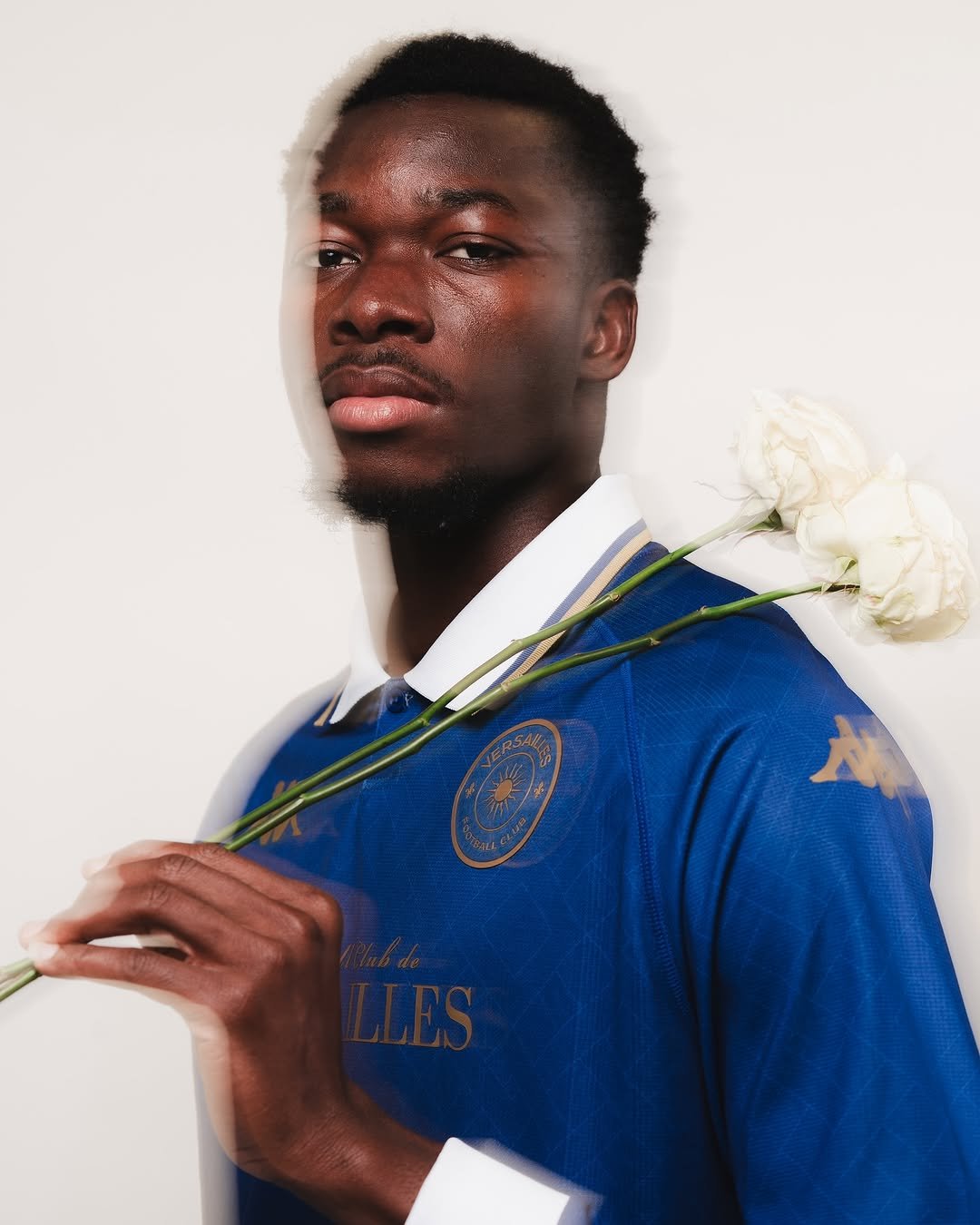Portrait of a young man with dark skin, wearing a blue sports jersey with a white collar, holding white flowers against a white background.