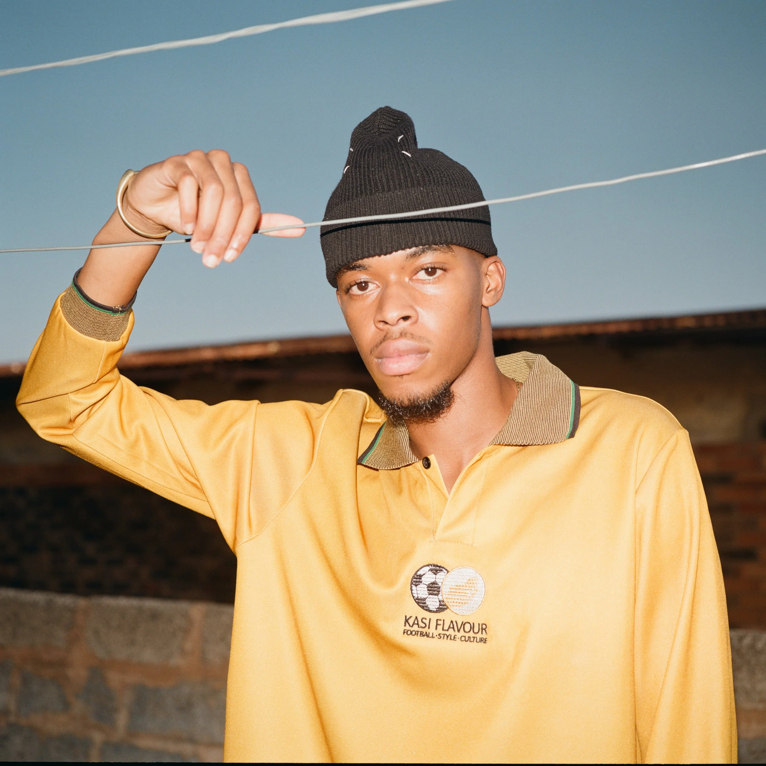 Young man wearing a black beanie, yellow long-sleeve shirt, and gold bracelet, holding a wire with his right hand, standing outdoors in front of a brick wall, with a clear sky in the background.