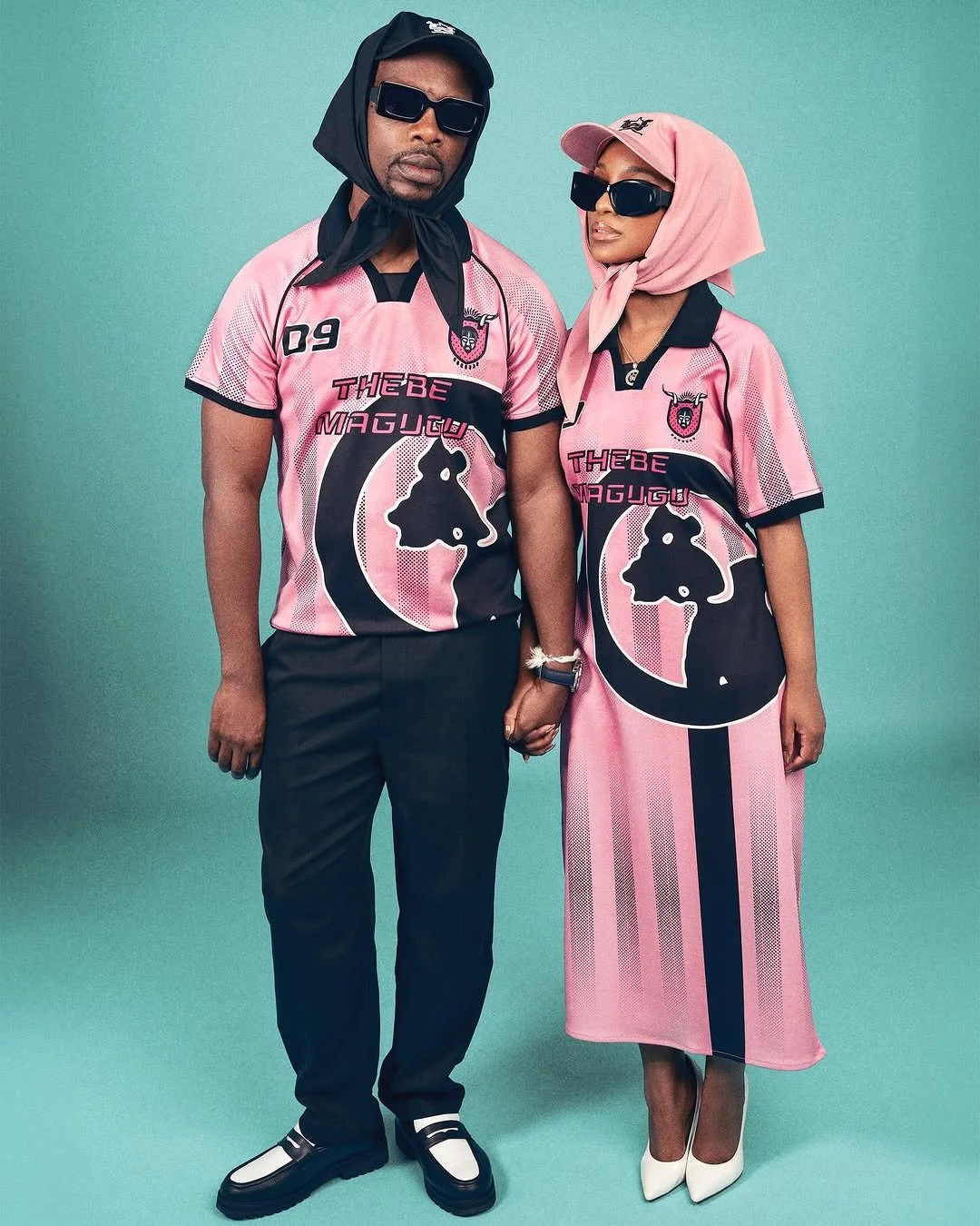 Two people standing against a teal background, holding hands, wearing matching pink and black sports jerseys with a bull logo and text, paired with stylish accessories and fashionable clothing items.