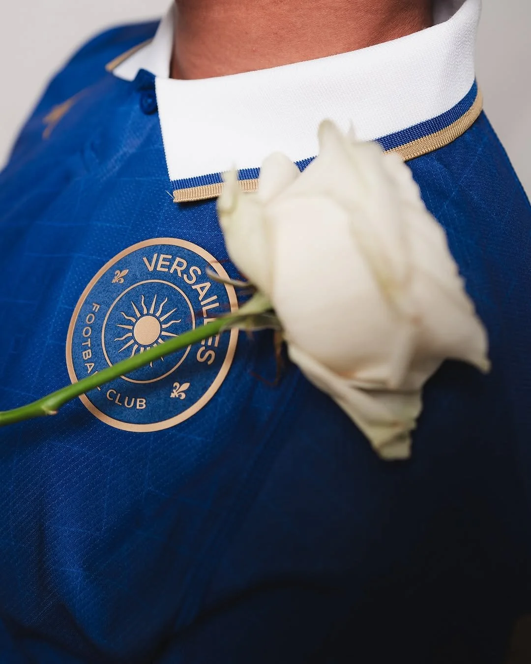 Person wearing a blue Versaires football club jersey with a white collar and holding a white rose.