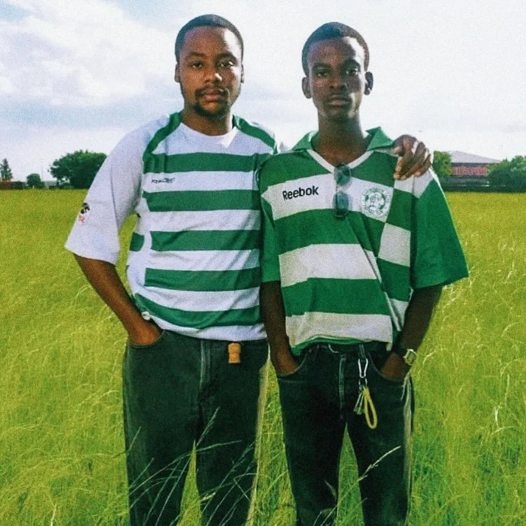 Two young men standing on a grassy field, wearing green and white striped sports jerseys, with one having sunglasses hanging from the collar. They have their arms around each other's shoulders.