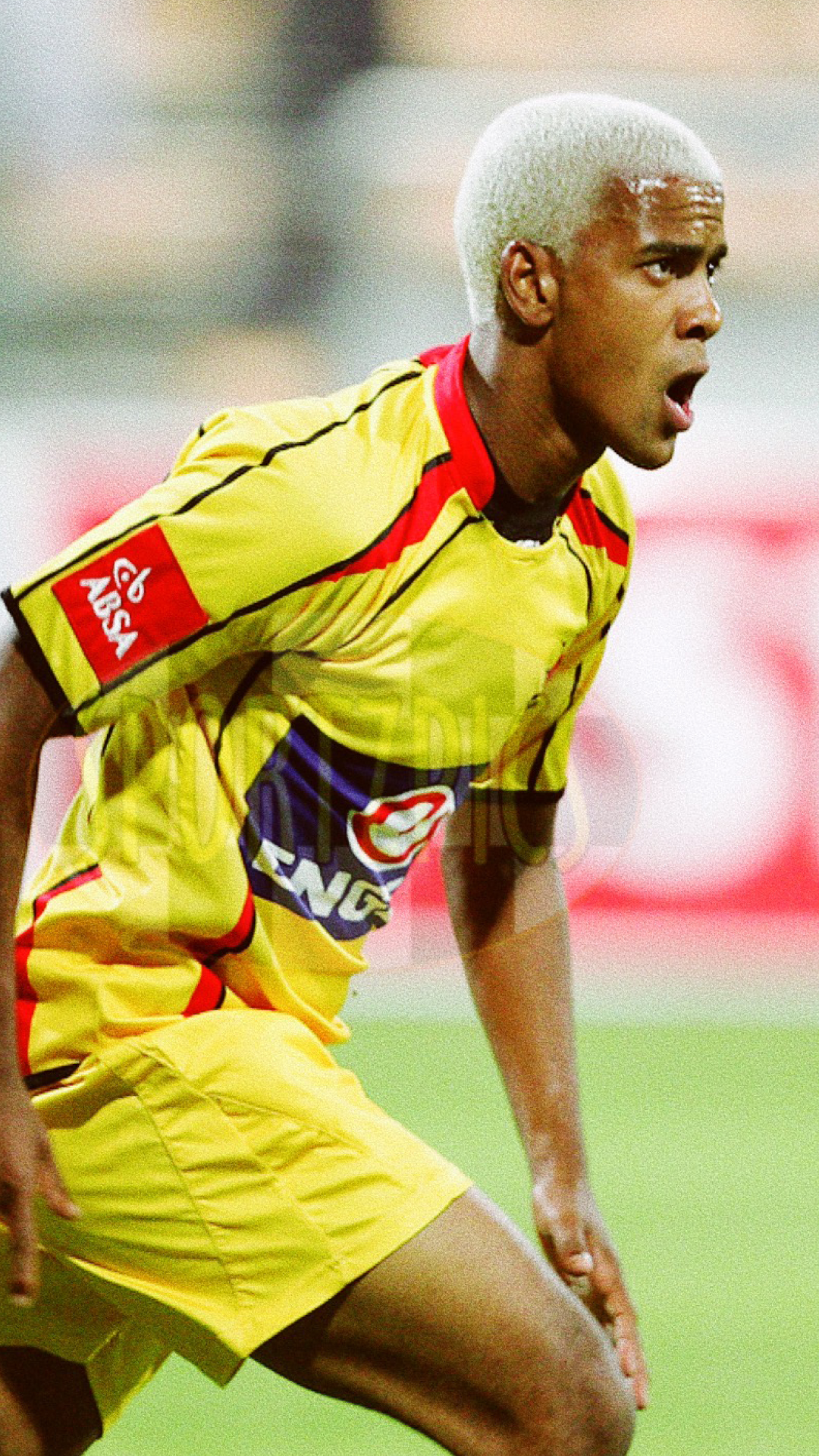 A soccer player in a yellow uniform with red and black accents, leaning forward with his mouth open, on a green field.