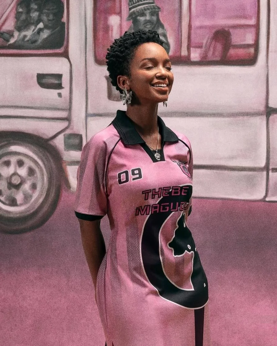 A smiling woman with short curly hair, wearing a pink and black sports jersey with the number 09 and the text "Thébe Magu" on it, standing in front of a pink background with a mural of a truck and a person.