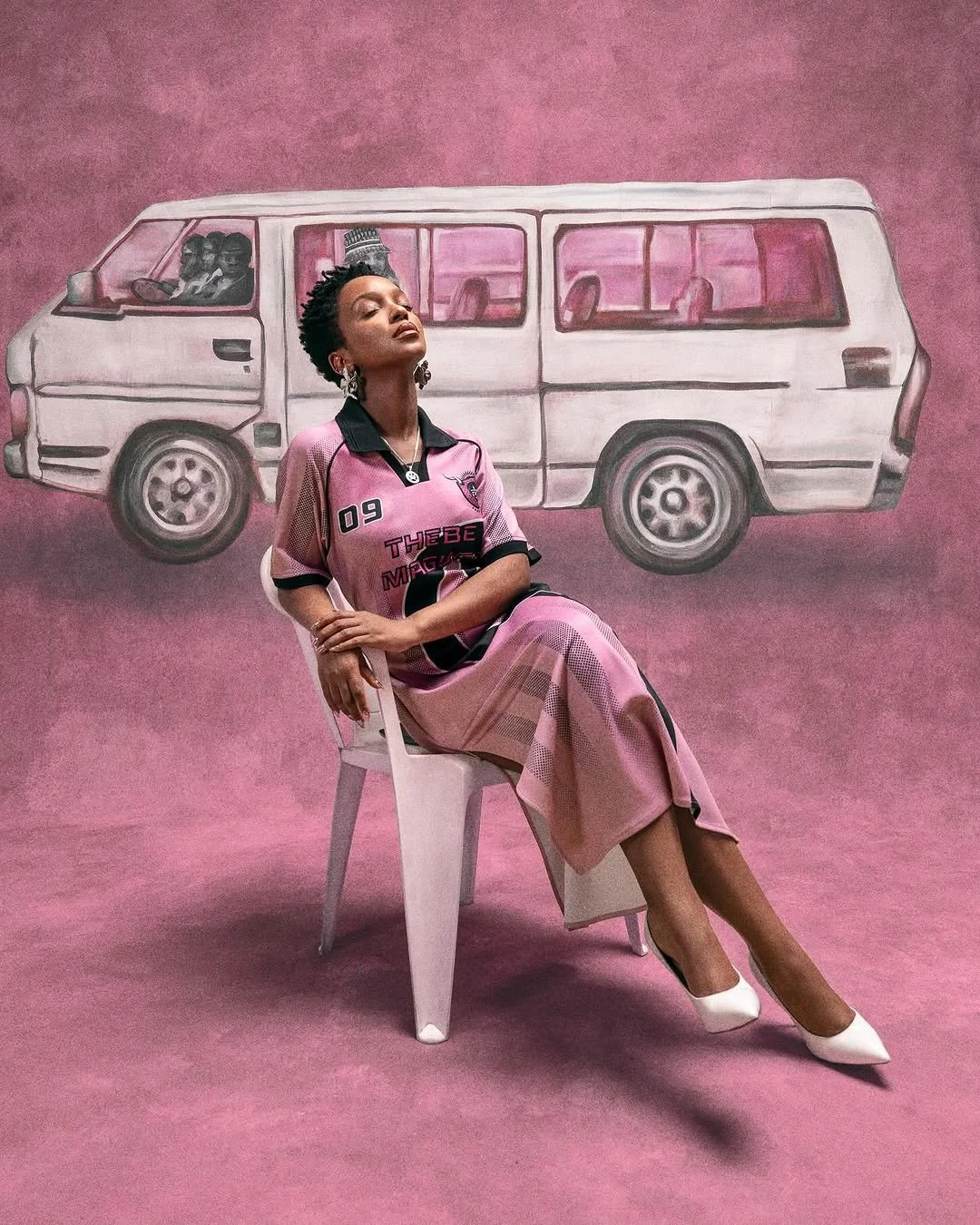 A woman in a pink sports dress sitting on a white chair with a relaxed pose, in front of a painted backdrop of a white van on a pink background.