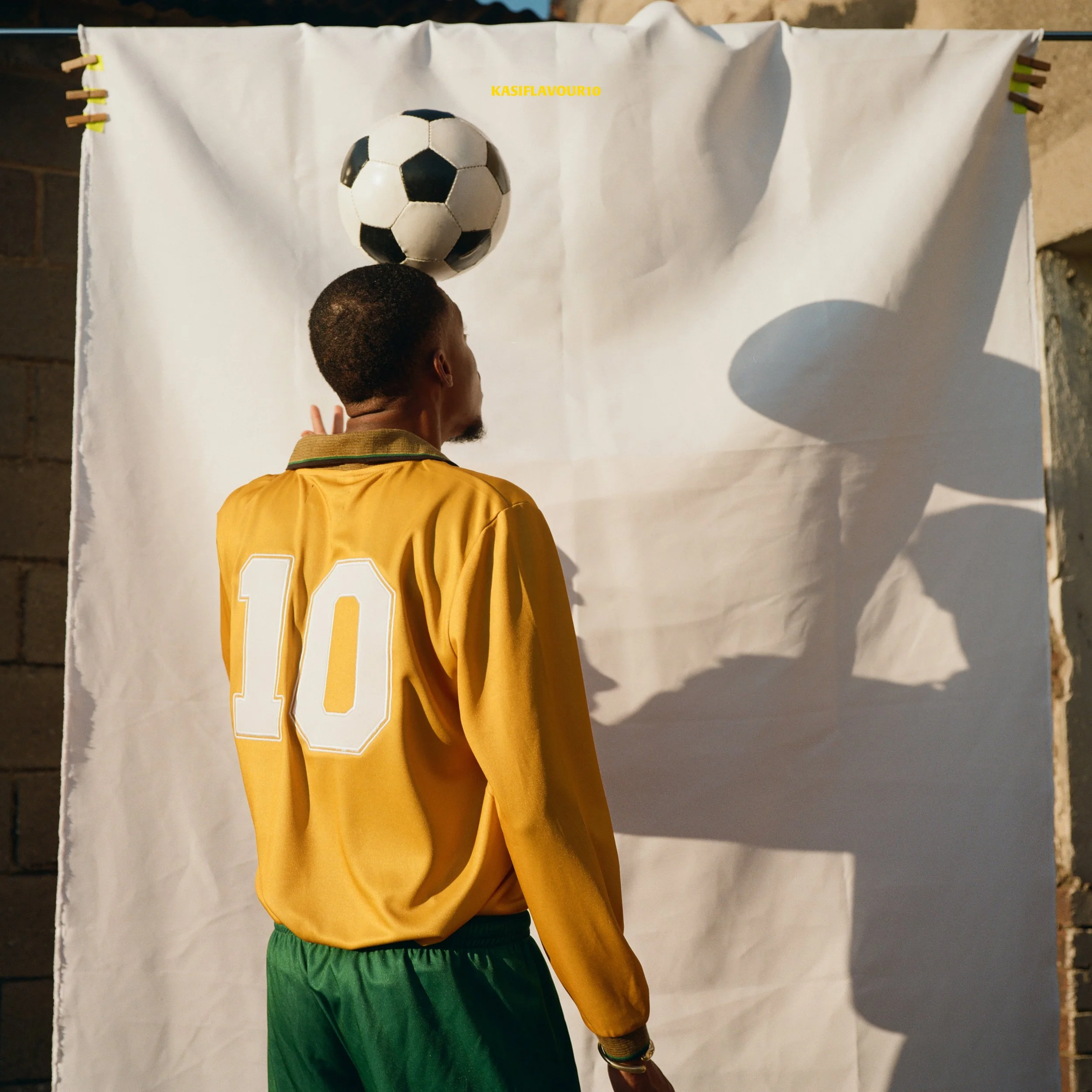 A man wearing a yellow sports jersey with the number 10 on the back, standing in front of a white backdrop, balancing a black and white soccer ball on his forehead. The backdrop has a shadow cast by the ball.