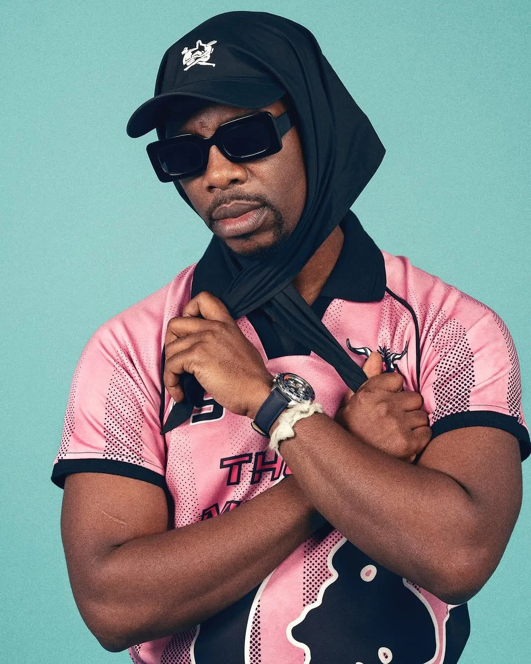 A man wearing a pink sports jersey, black sunglasses, a black cap, and a watch, with a scarf wrapped around his head and neck, standing against a light blue background.