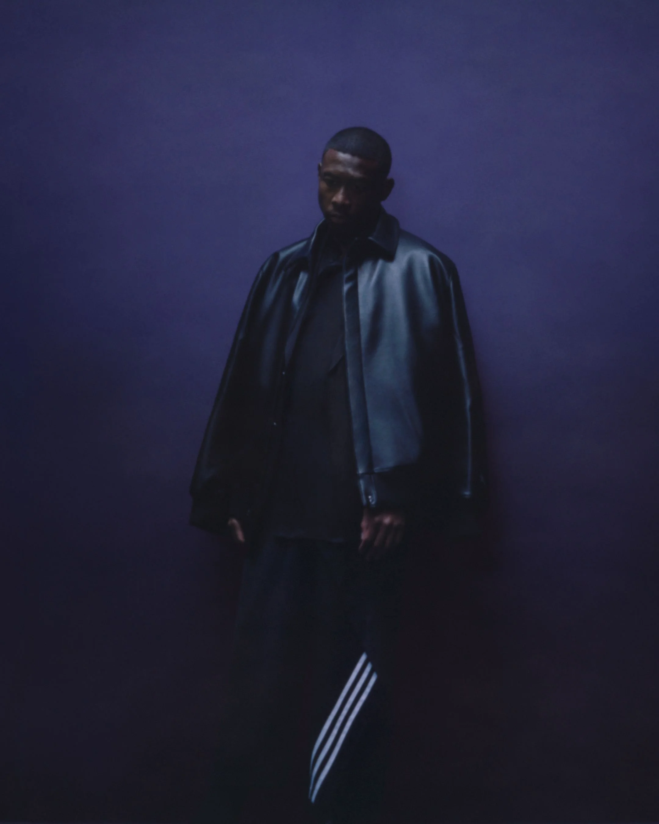 Young man in a black leather jacket and black clothing, standing against a dark purple background.
