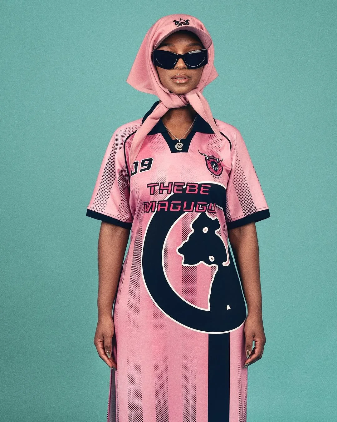 A woman wearing a pink sports jersey with black and white accents, a pink headscarf, black sunglasses, and a cap, standing against a teal background.